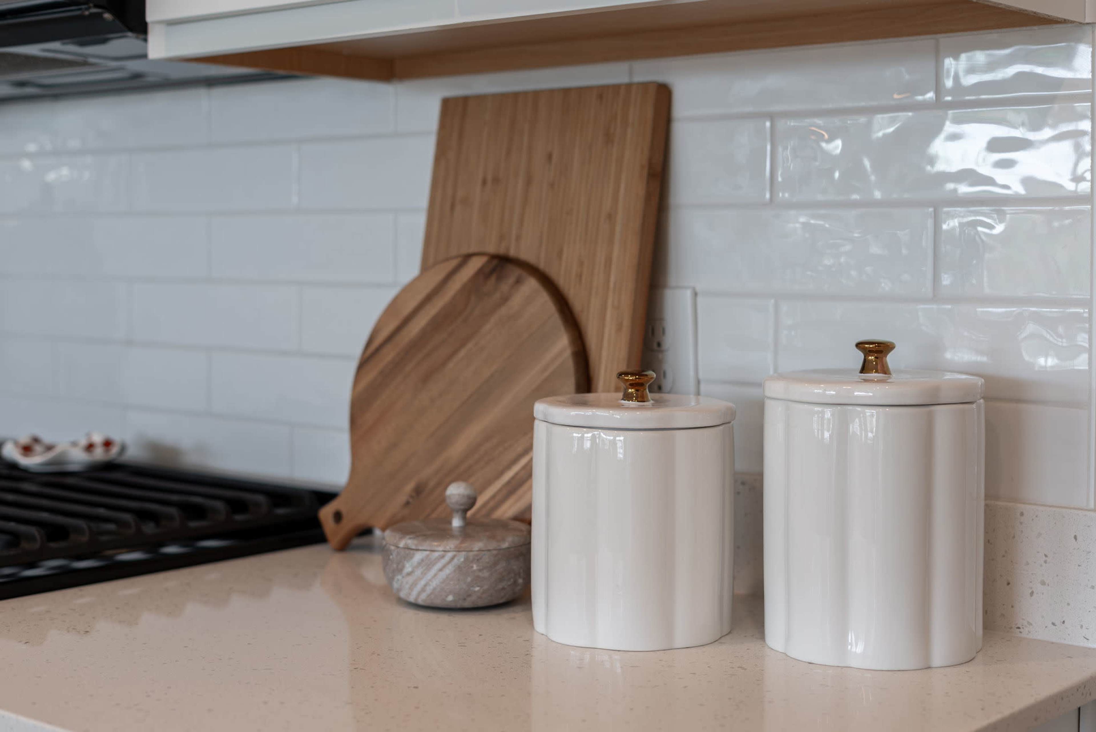 The image shows a kitchen countertop with two white canisters, a round wooden cutting board, a small lidded glass container, and a stove in the background.
