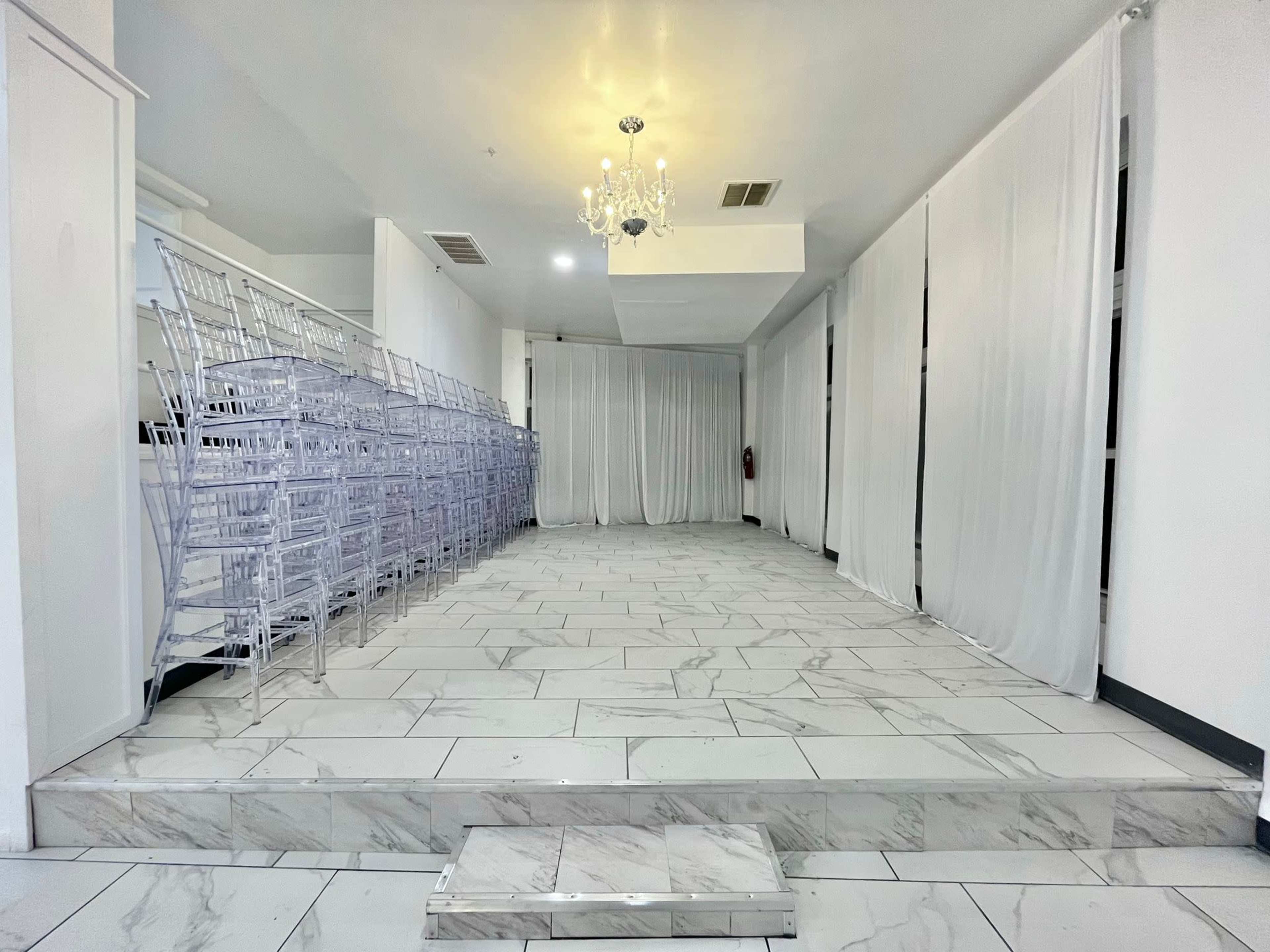 A spacious, well-lit room with white drapes, marble flooring, and stacked transparent Chiavari chairs along one side.
