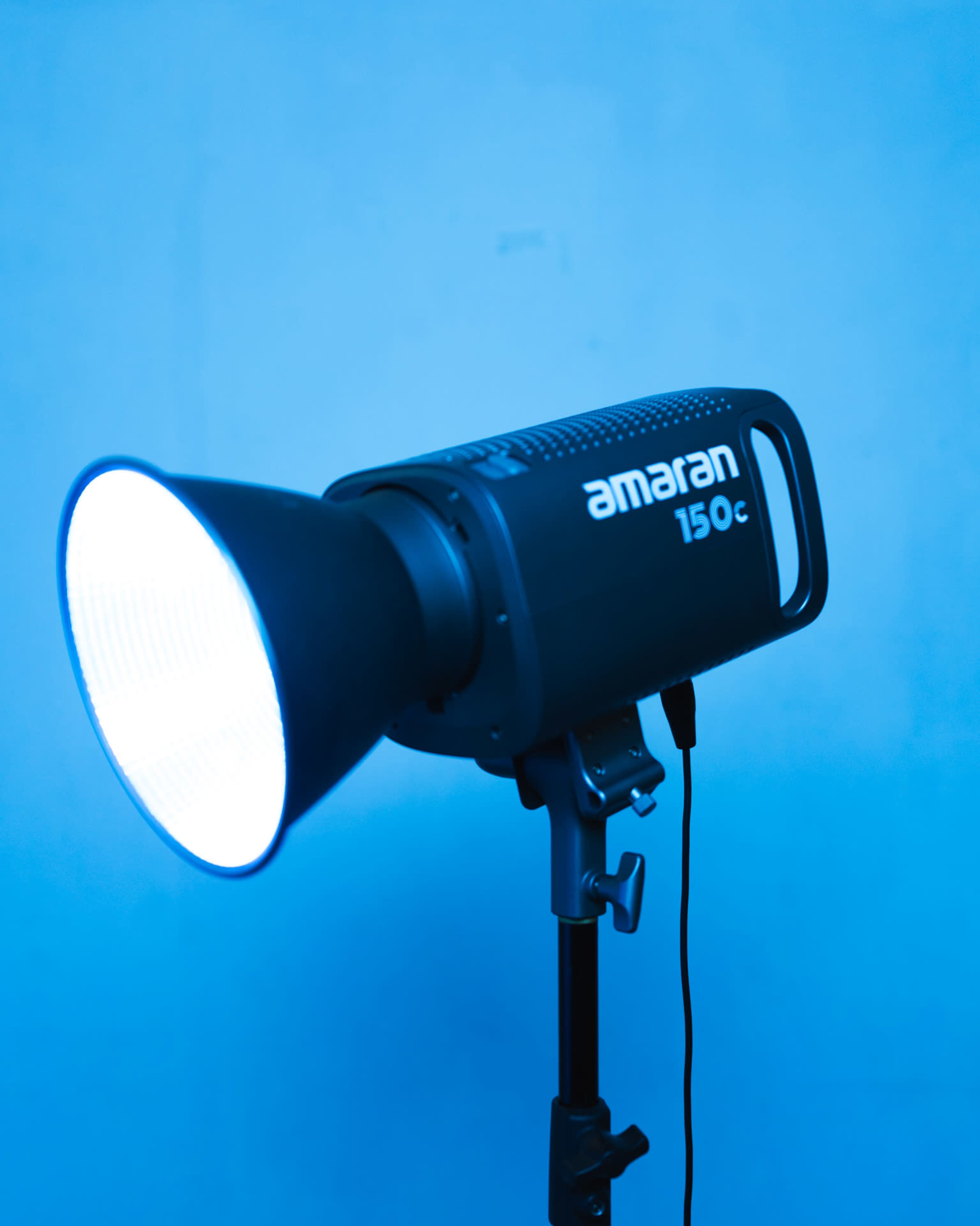 A black Amaran 150c studio light on a stand, emitting a blue-tinted light.