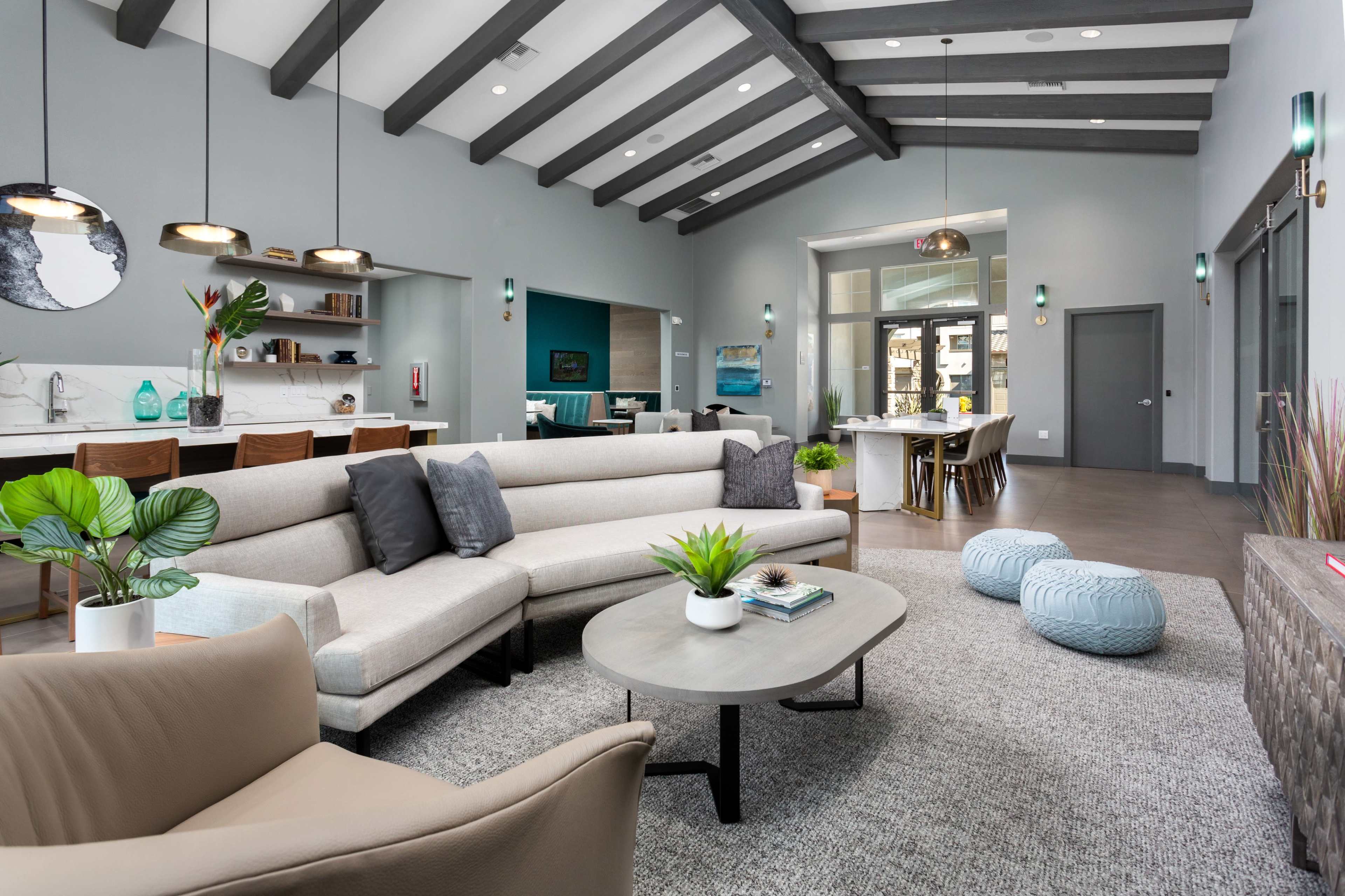 A modern living space features a sectional sofa, a coffee table, and a dining area with a wooden table and blue stools, surrounded by light gray walls and exposed wooden beams.