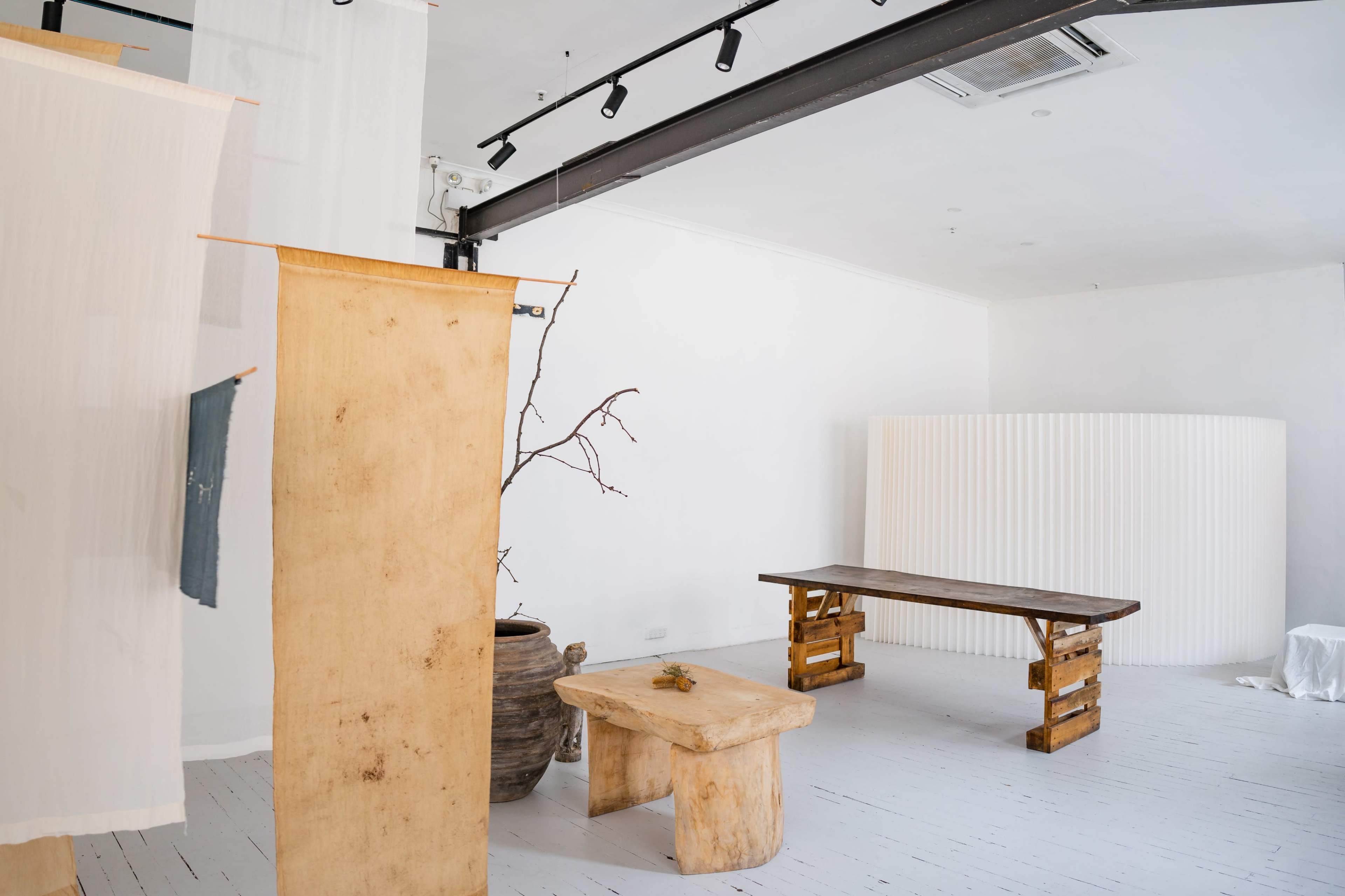 The image features a minimalistic interior scene with wooden furniture, a textured wall panel, and decorative elements like a branch and clay pot.