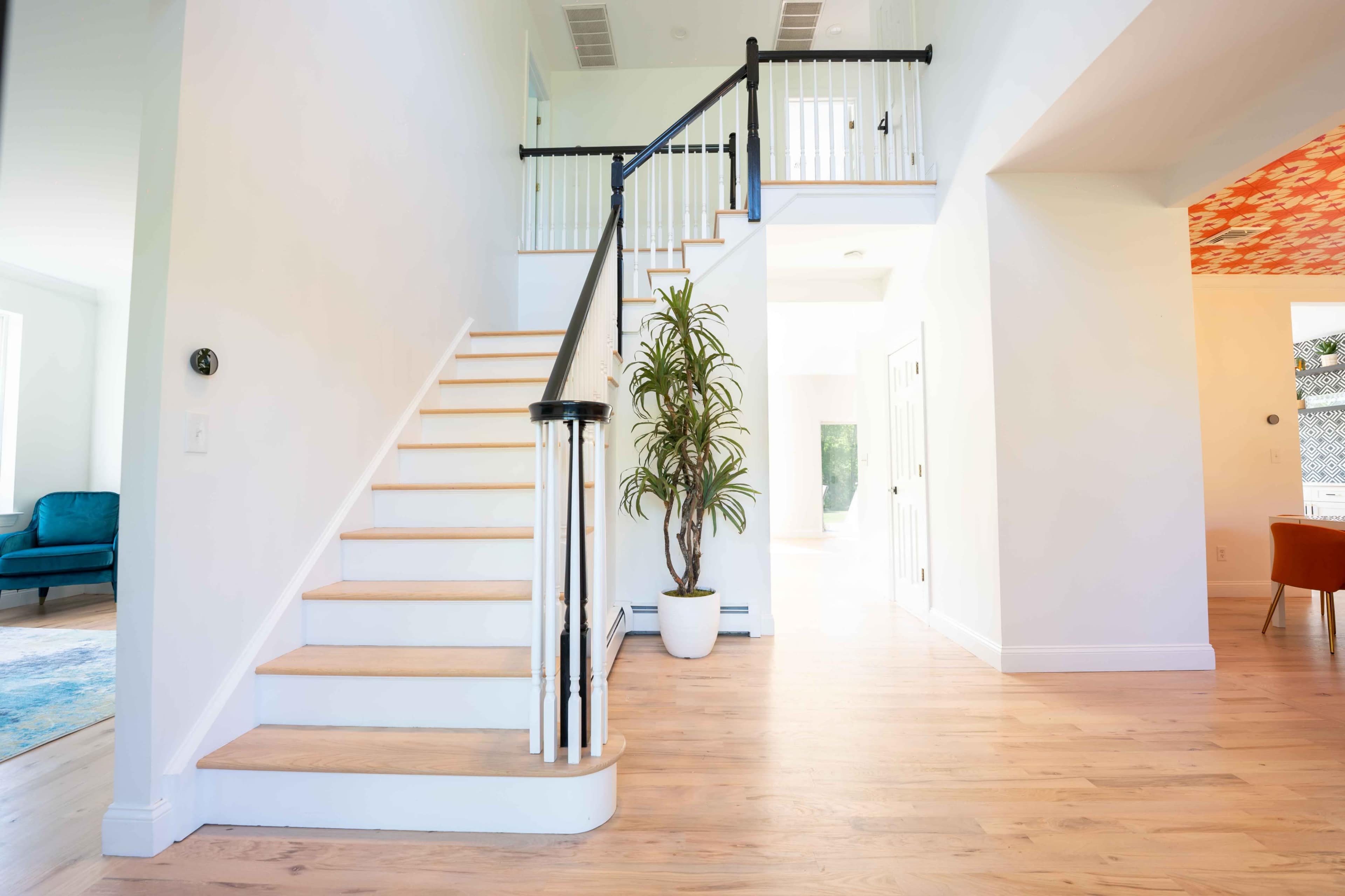 A staircase with light wood steps and black railings leads to an upstairs landing in a brightly lit foyer with a potted plant.