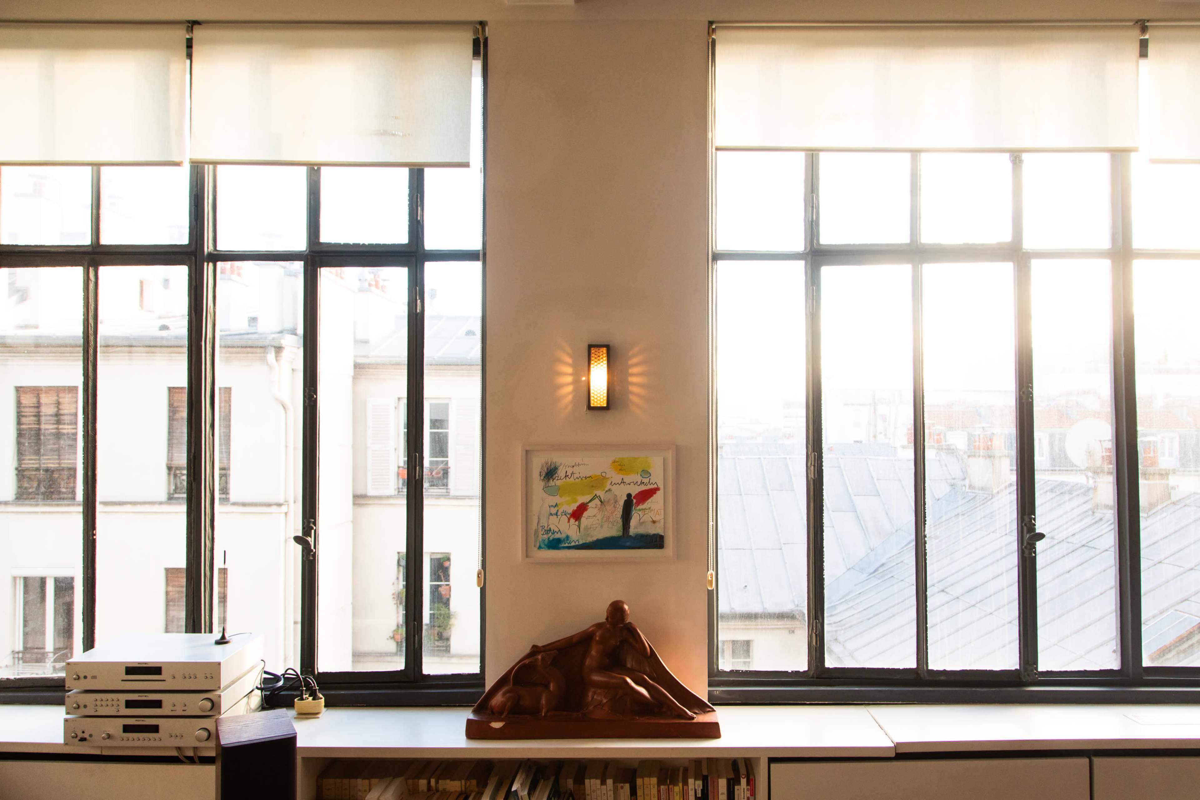 The image shows a well-lit interior space with large windows revealing a view of buildings outside, featuring a piece of artwork on the wall and a wooden sculpture on a shelf.