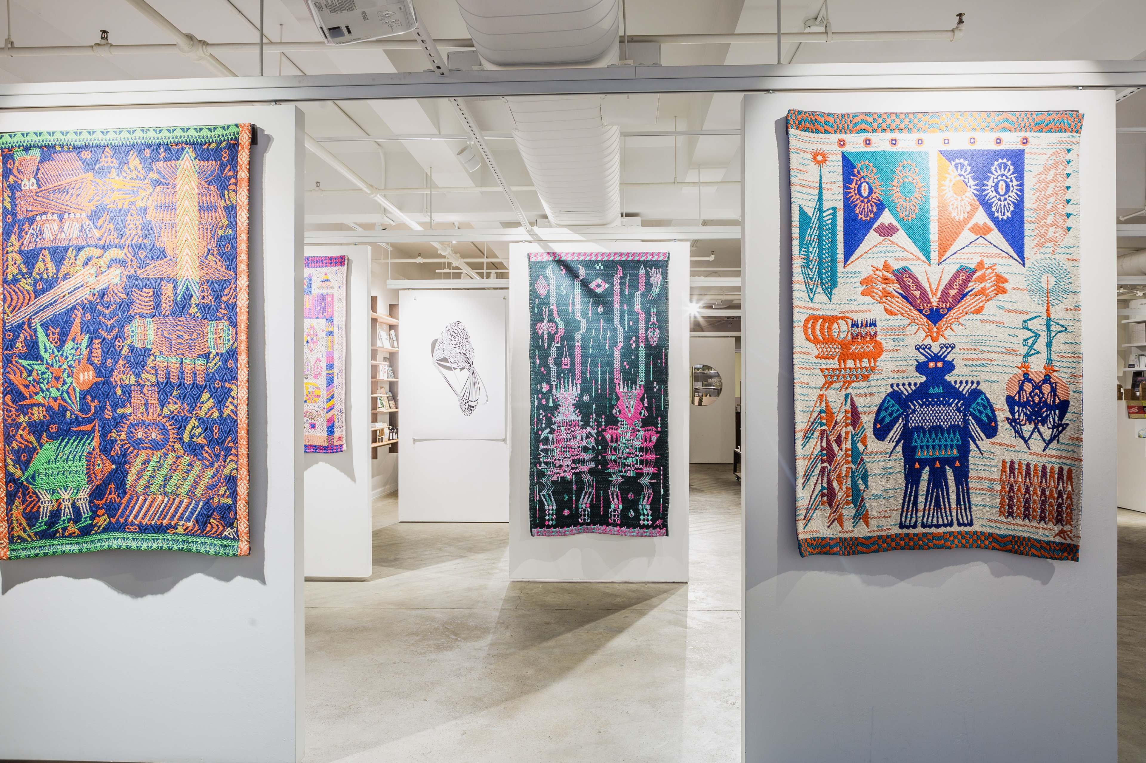 A gallery space featuring colorful textile artworks hanging on white walls, with a central illustration visible in the background.