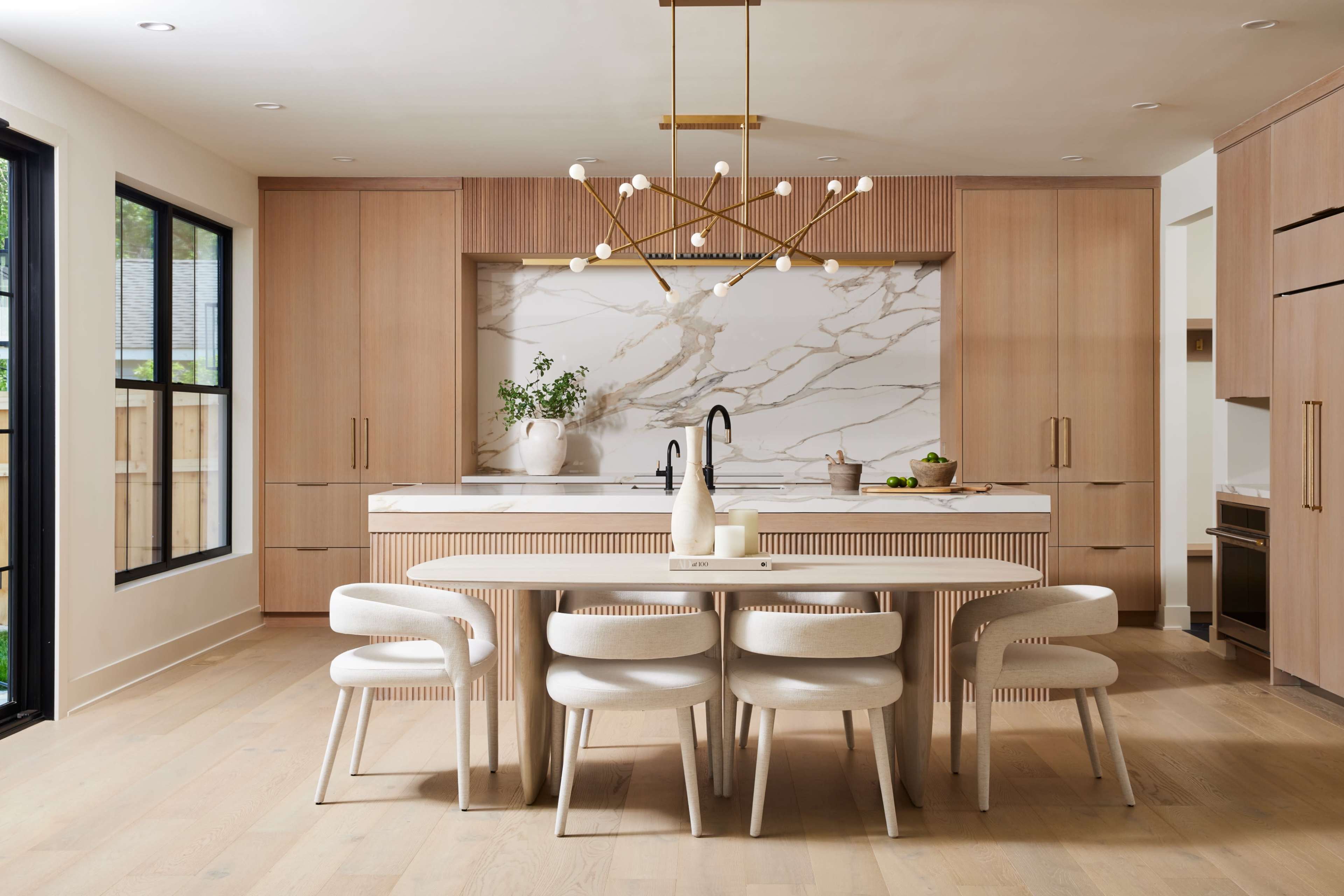 A modern kitchen features light wooden cabinetry, a marble backsplash, and a dining area with a round table and upholstered chairs.