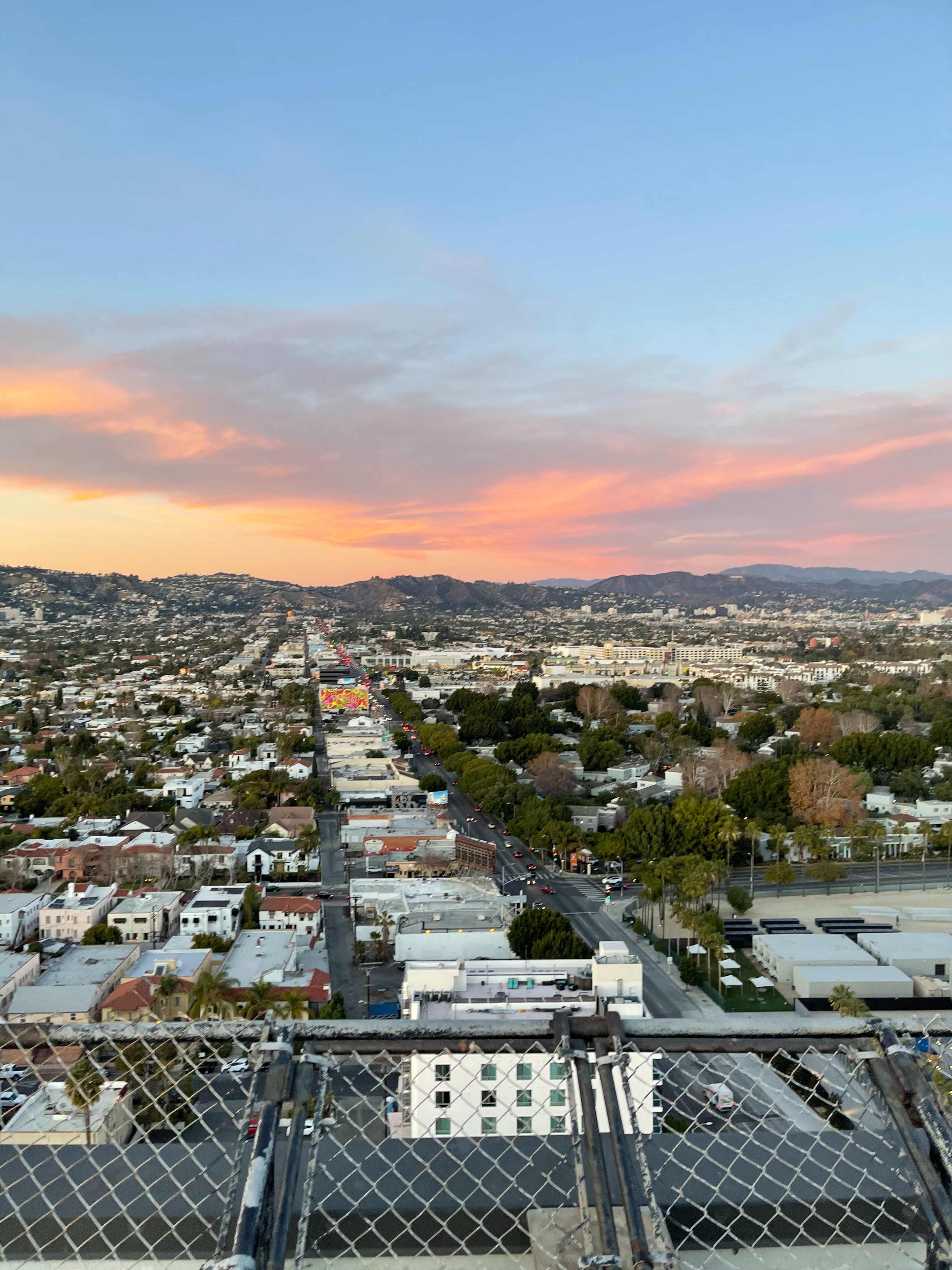 A cityscape at sunset with streets lined by buildings and distant hills under a colorful sky.