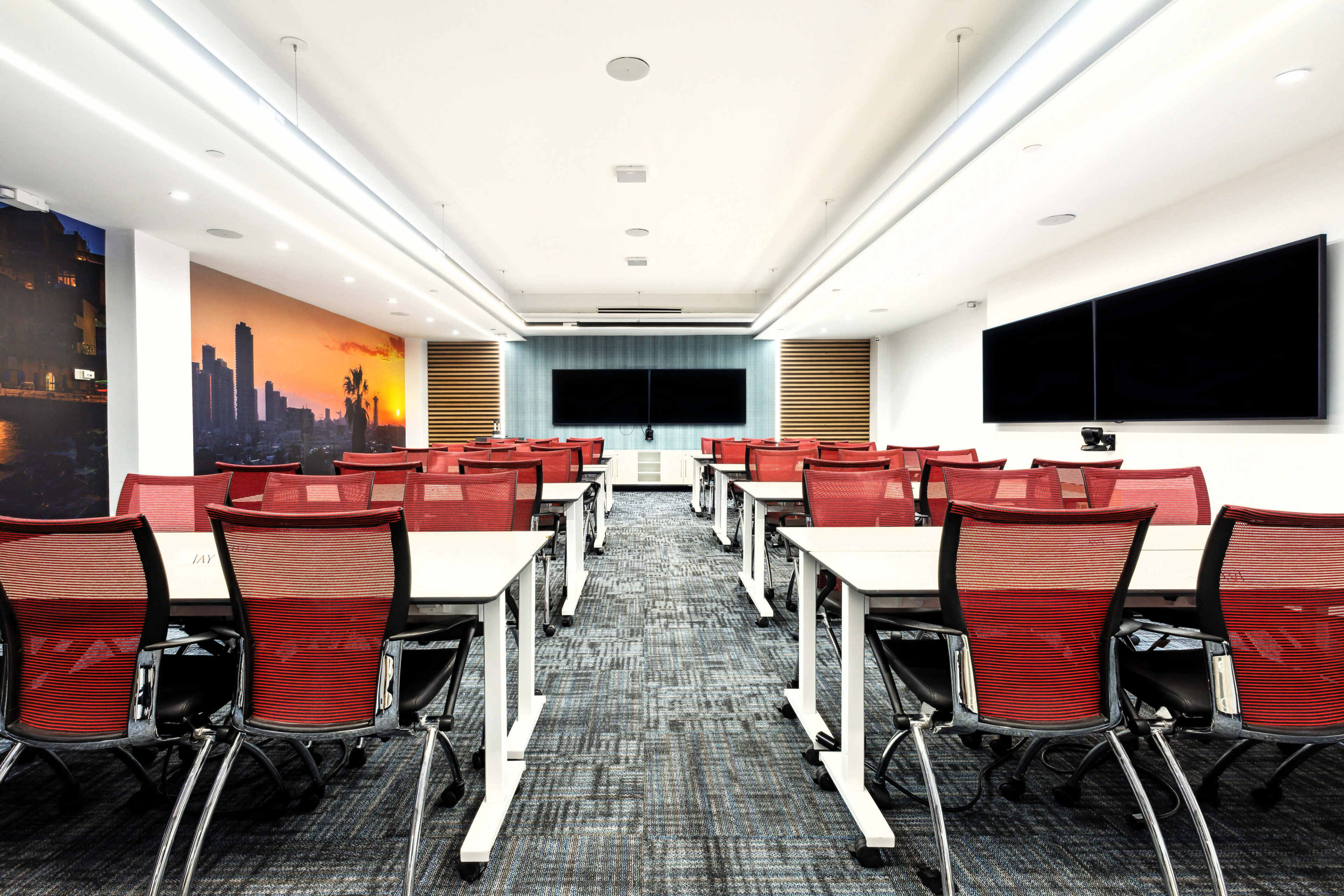 The image shows a modern conference room set up with multiple rows of tables and red chairs, featuring two large screens at the front and a cityscape mural on one wall.