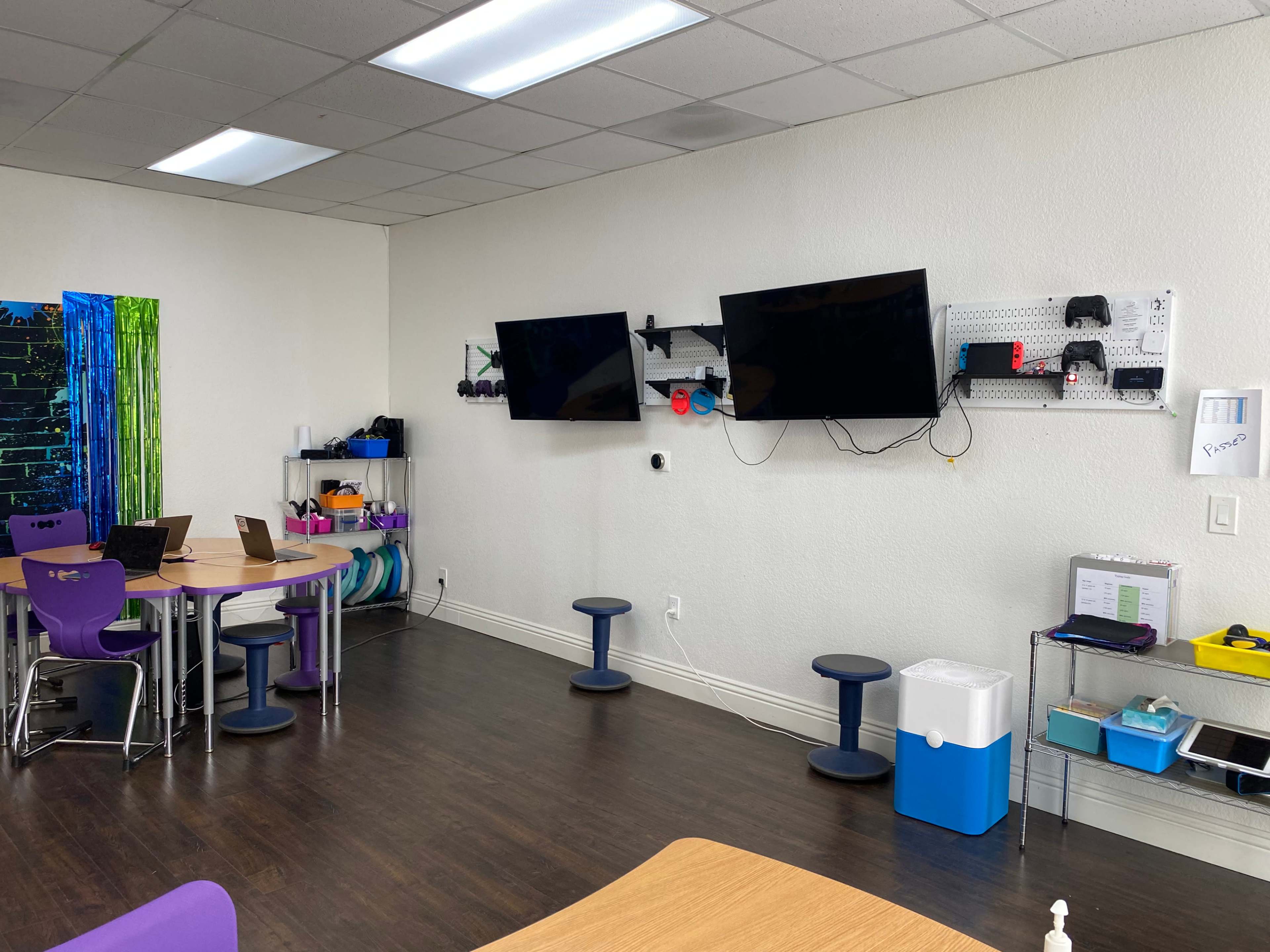 The image shows a classroom or activity space with two mounted TVs, various seating options, and shelves displaying educational materials and equipment.