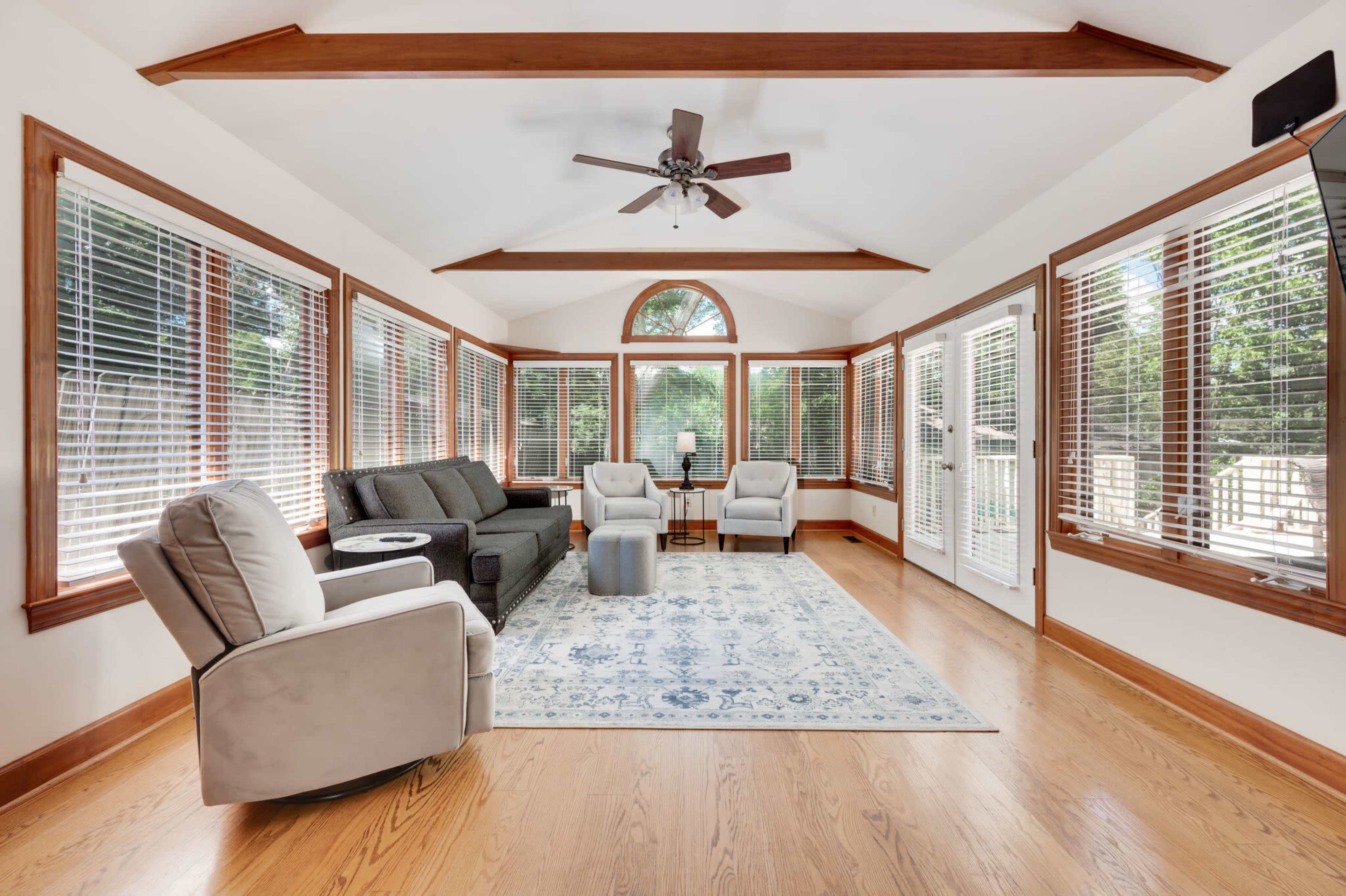 A sunroom features large windows with wooden frames, two armchairs, a sofa, and a decorative rug on wooden flooring.
