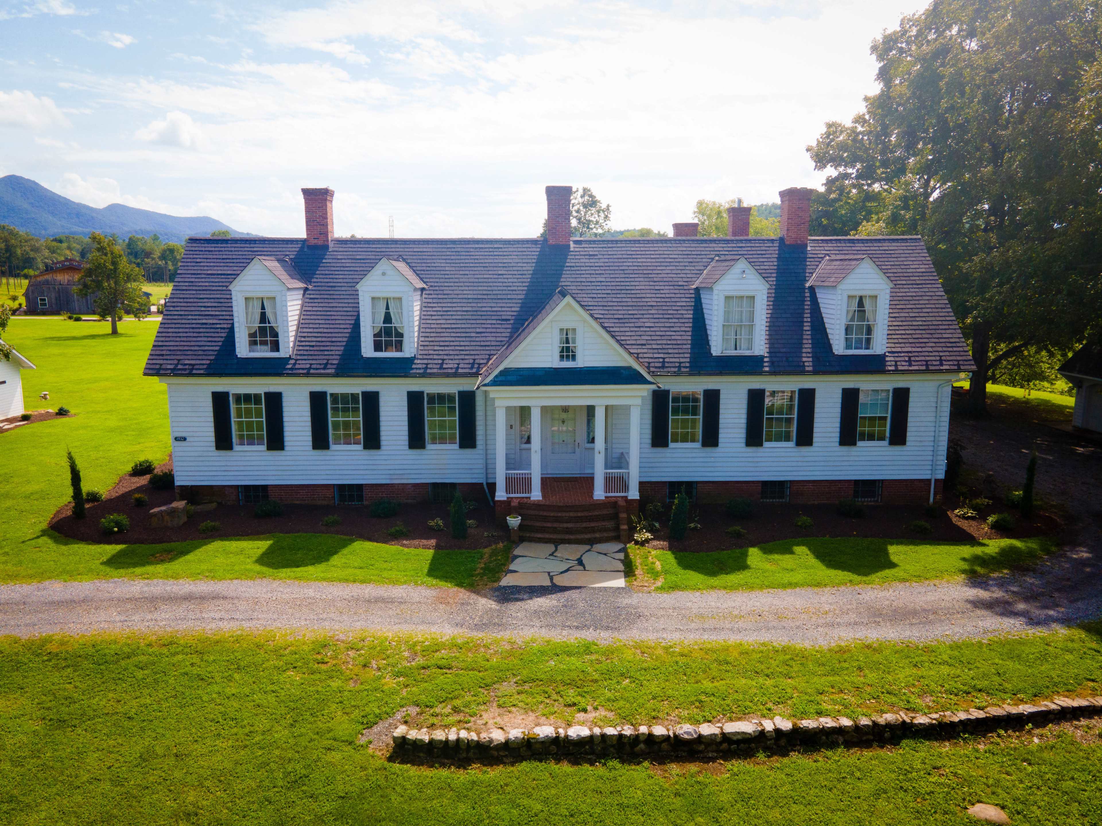 A large, two-story house with a symmetrical facade and a stone pathway leading to the front door is situated on a grassy landscape with trees in the background.
