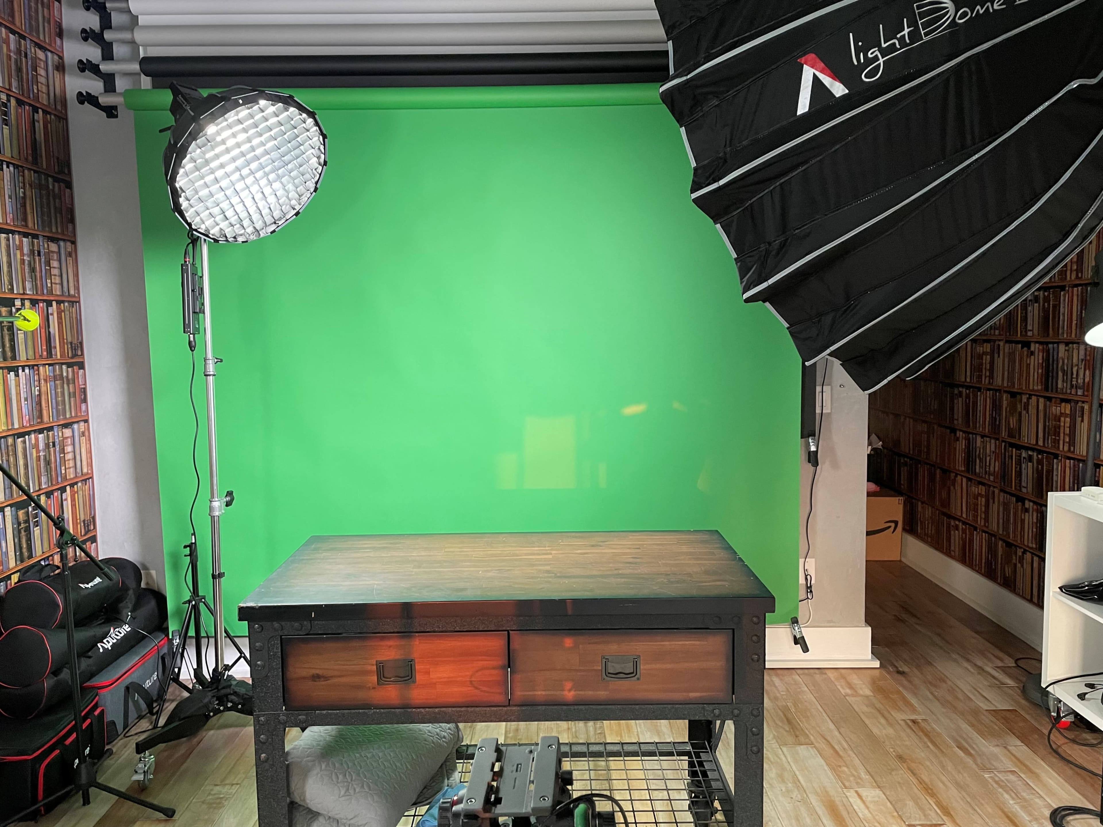 The image shows a studio setup featuring a green screen, a large softbox light, and a wooden table in front of a bookshelf.