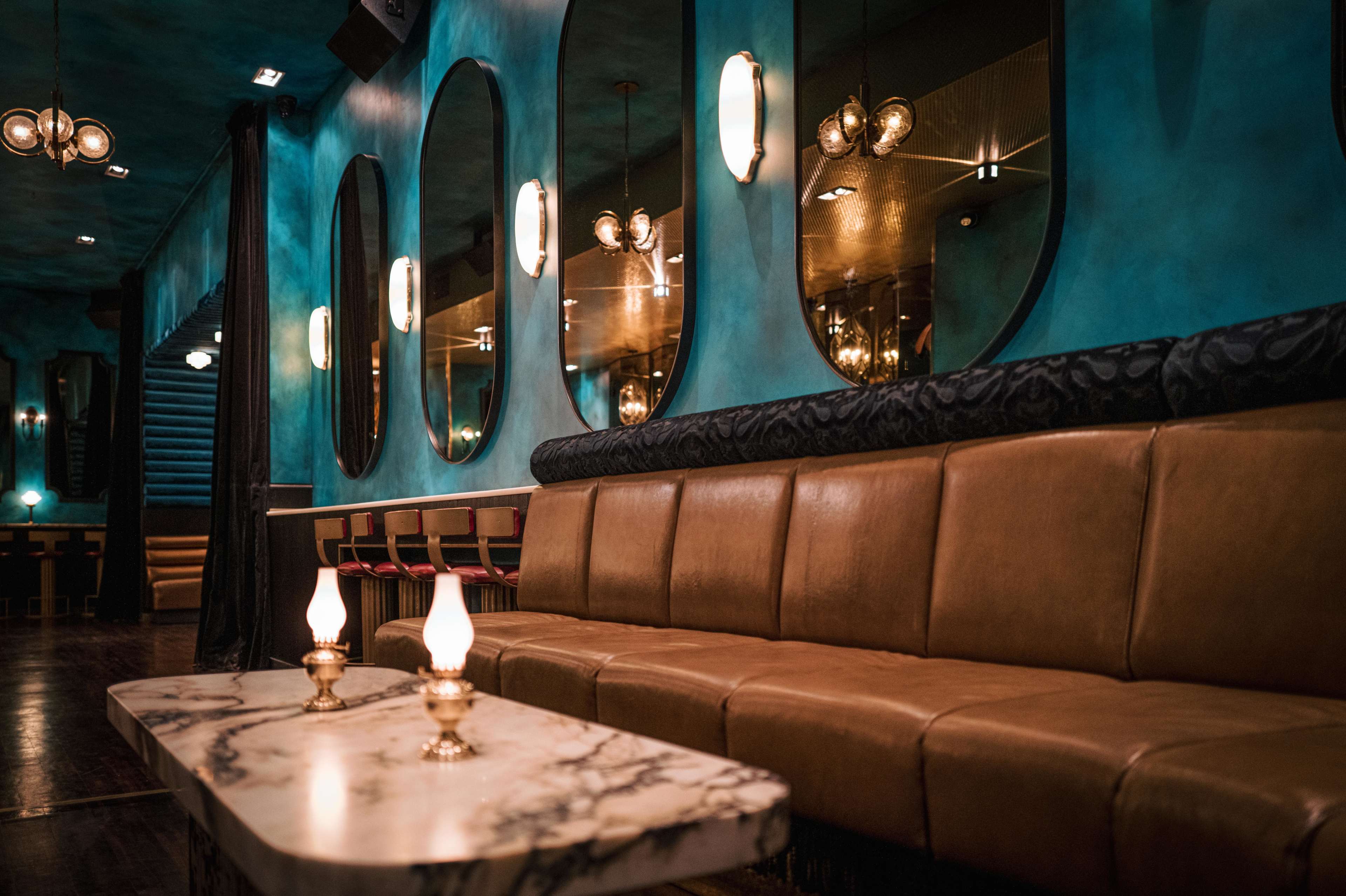 The image shows a stylish lounge area featuring curved mirrors, a brown tufted bench, and a marble table with small candle holders.