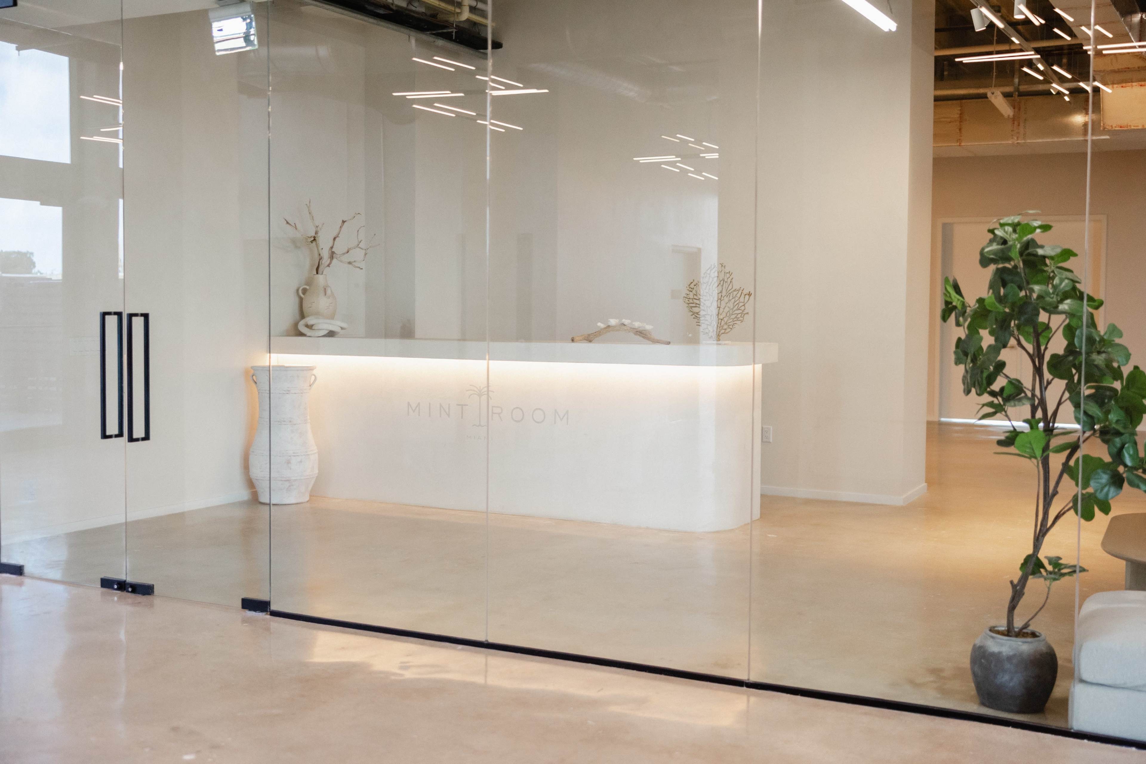 The image shows a sleek reception area with a white counter labeled "MINT ROOM," framed by large glass walls and minimalistic decor.