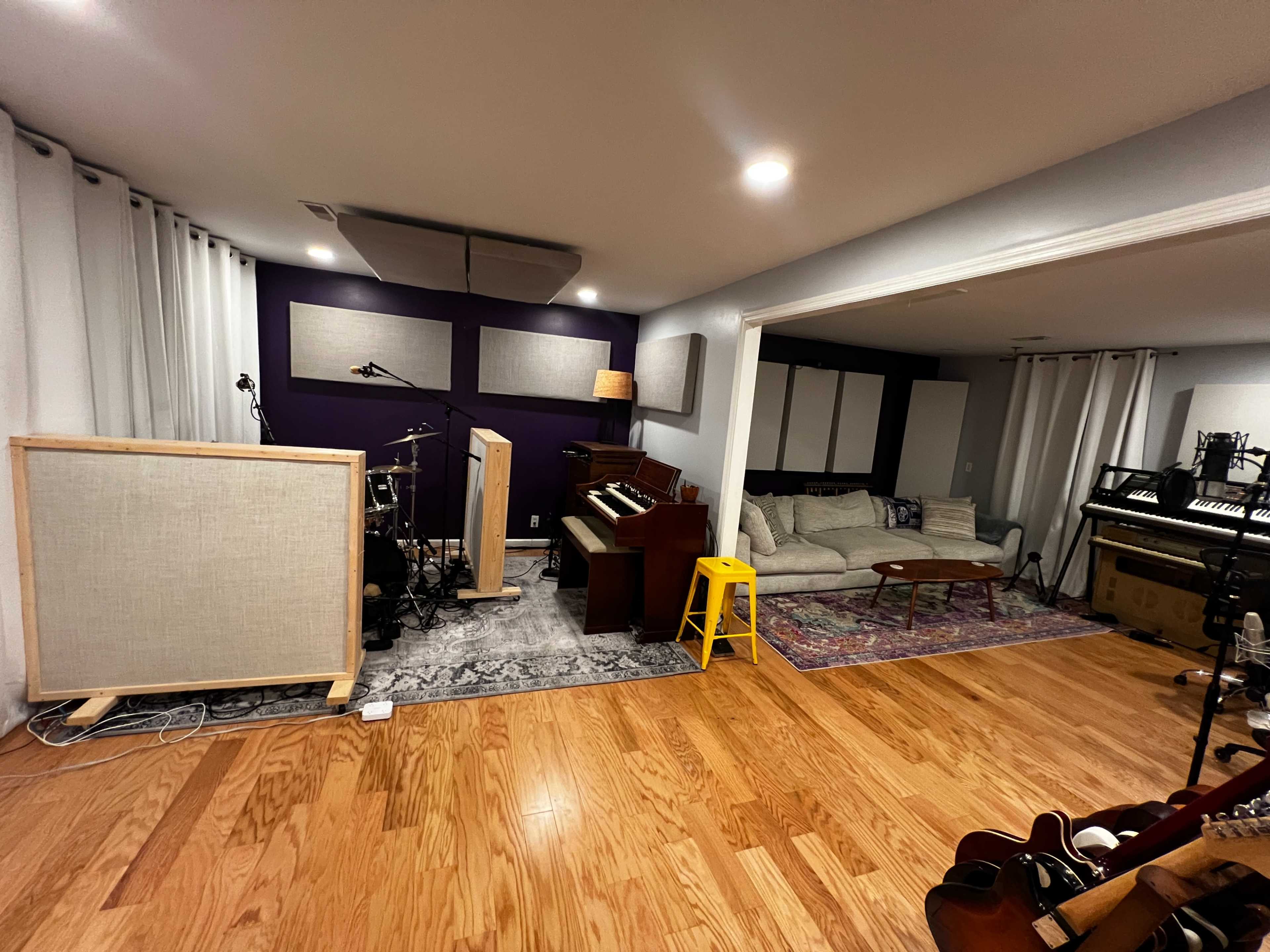 The image depicts a spacious music studio with a drum set, an electric piano, a comfortable couch, and various musical instruments arranged across a hardwood floor.