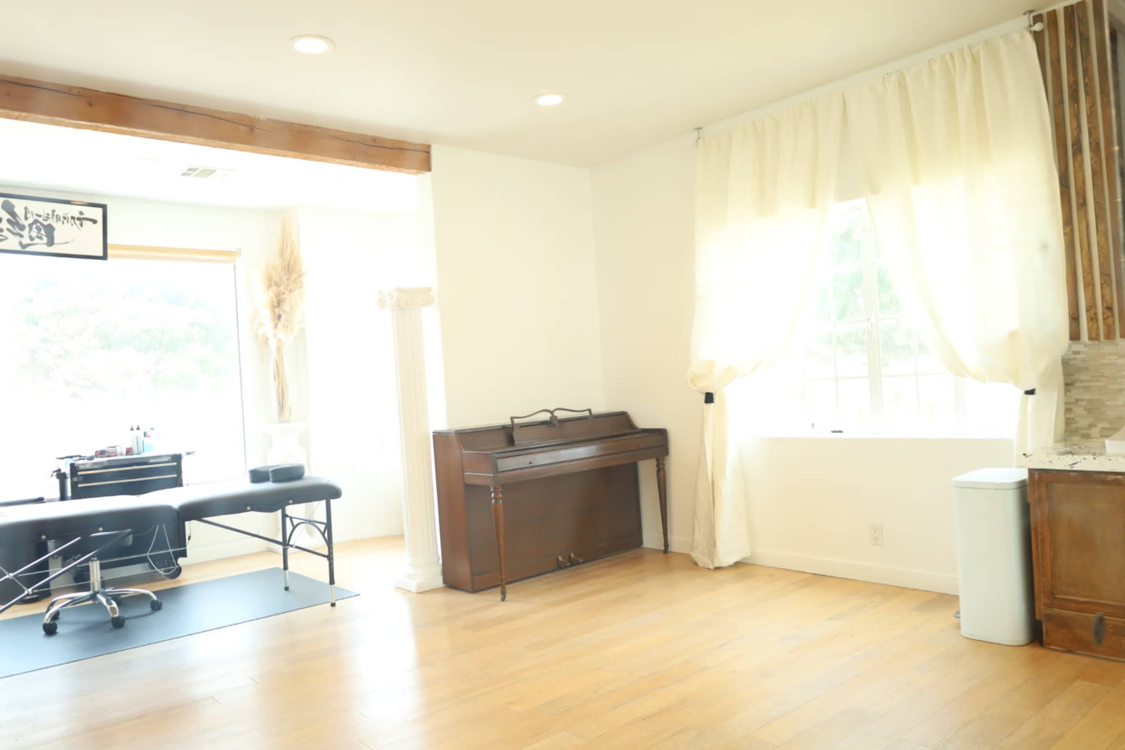The image shows a bright room featuring a piano, a treatment table, large windows with curtains, and wooden flooring.