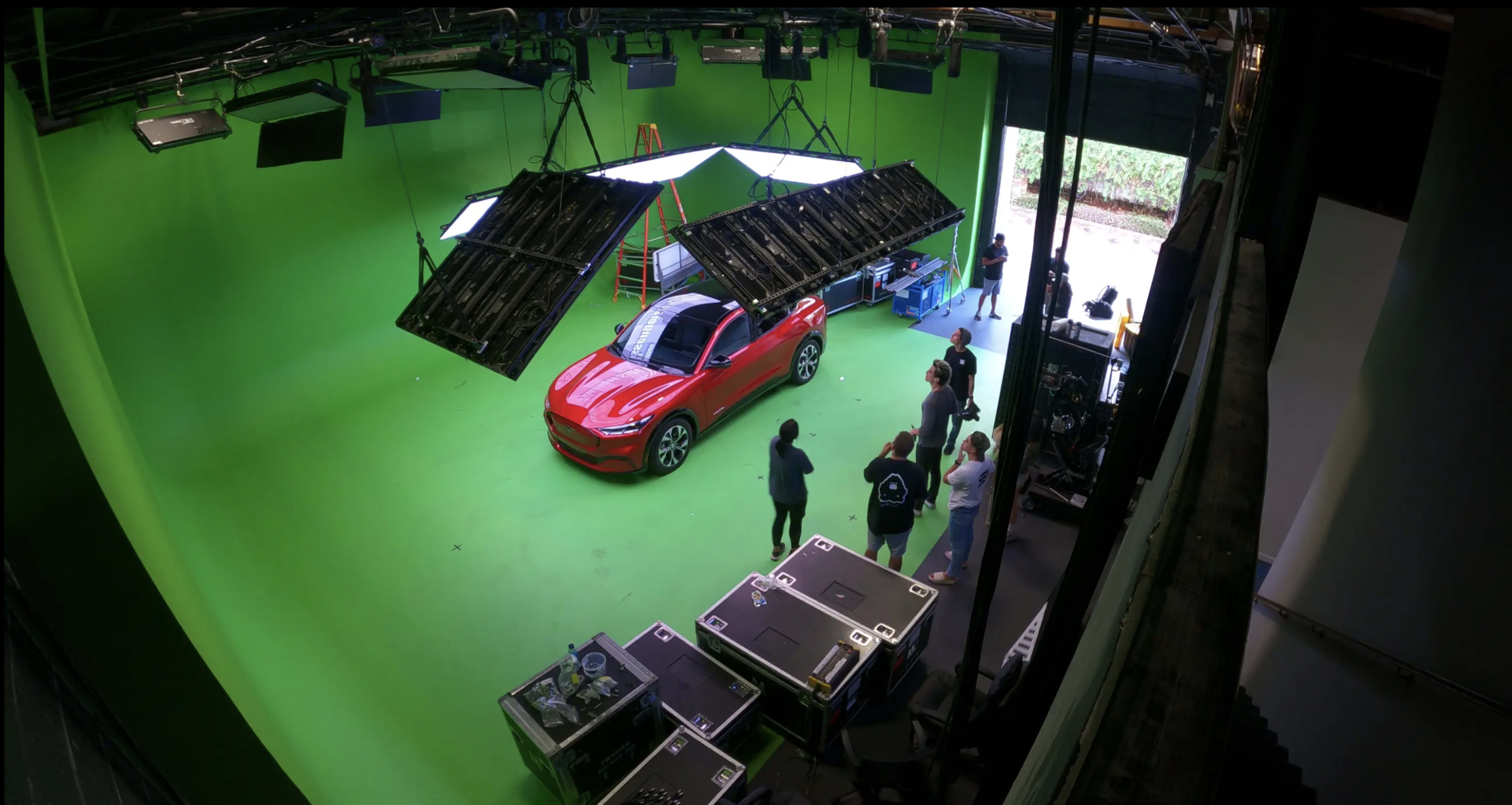A red car is positioned on a green screen stage with crew members and equipment in the background.