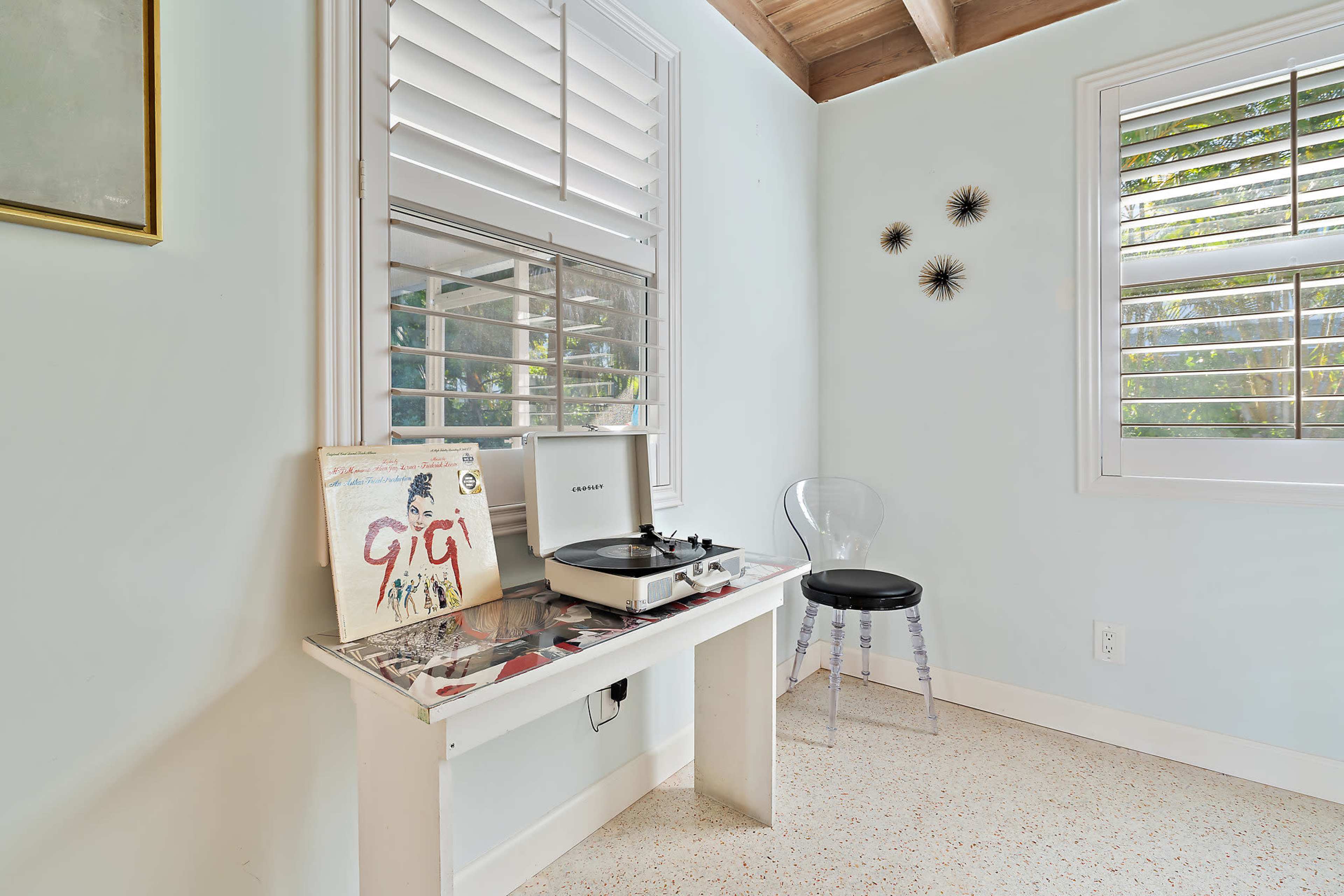 A record player and a vinyl album are placed on a white table beside a clear acrylic chair in a softly lit room with shuttered windows.