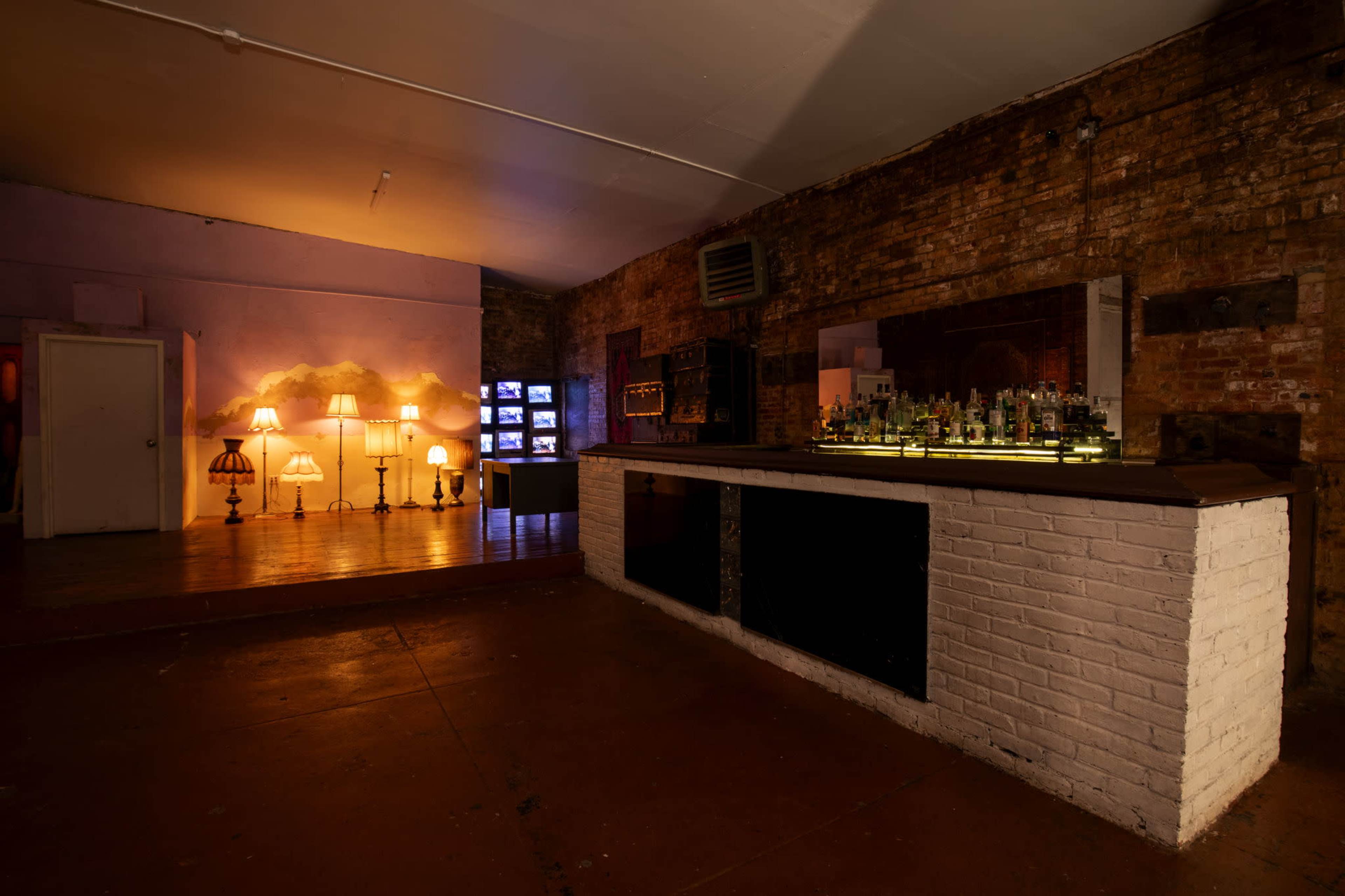 The image shows an indoor bar area with a white-brick counter, a collection of bottles on display, and ambient lighting from several standing lamps.