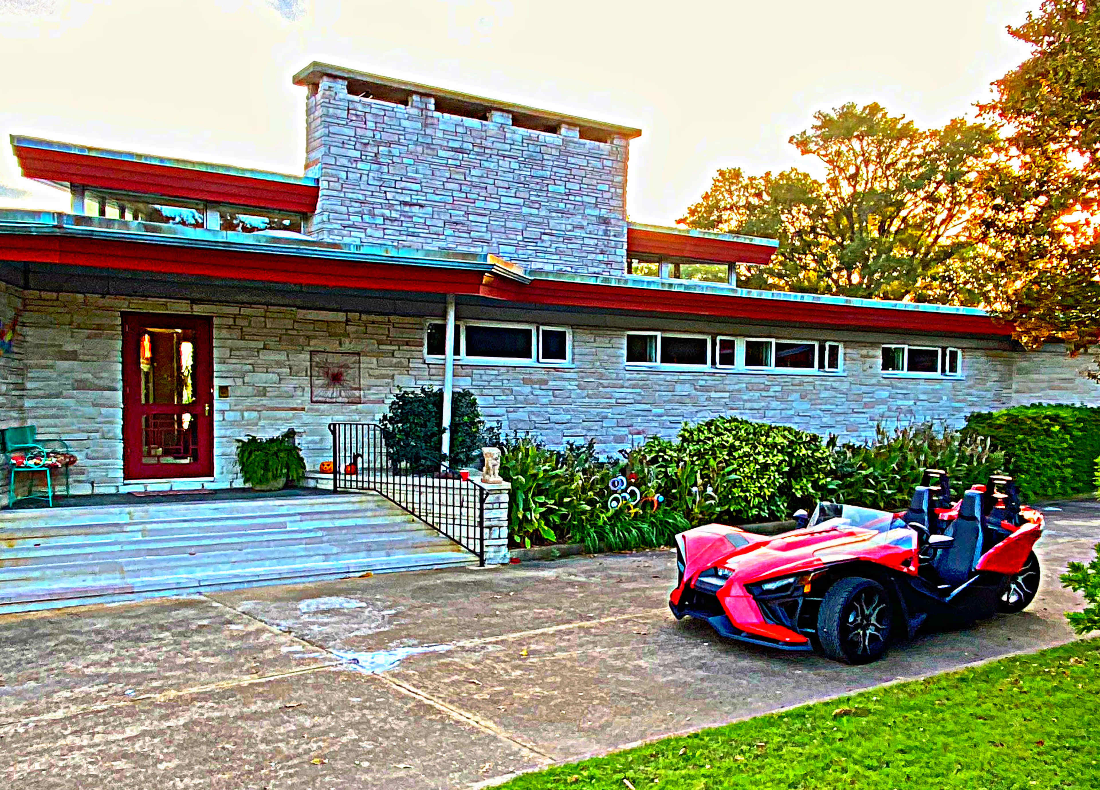 A red three-wheeled vehicle is parked in front of a modern stone house with a flat roof and landscaped garden.
