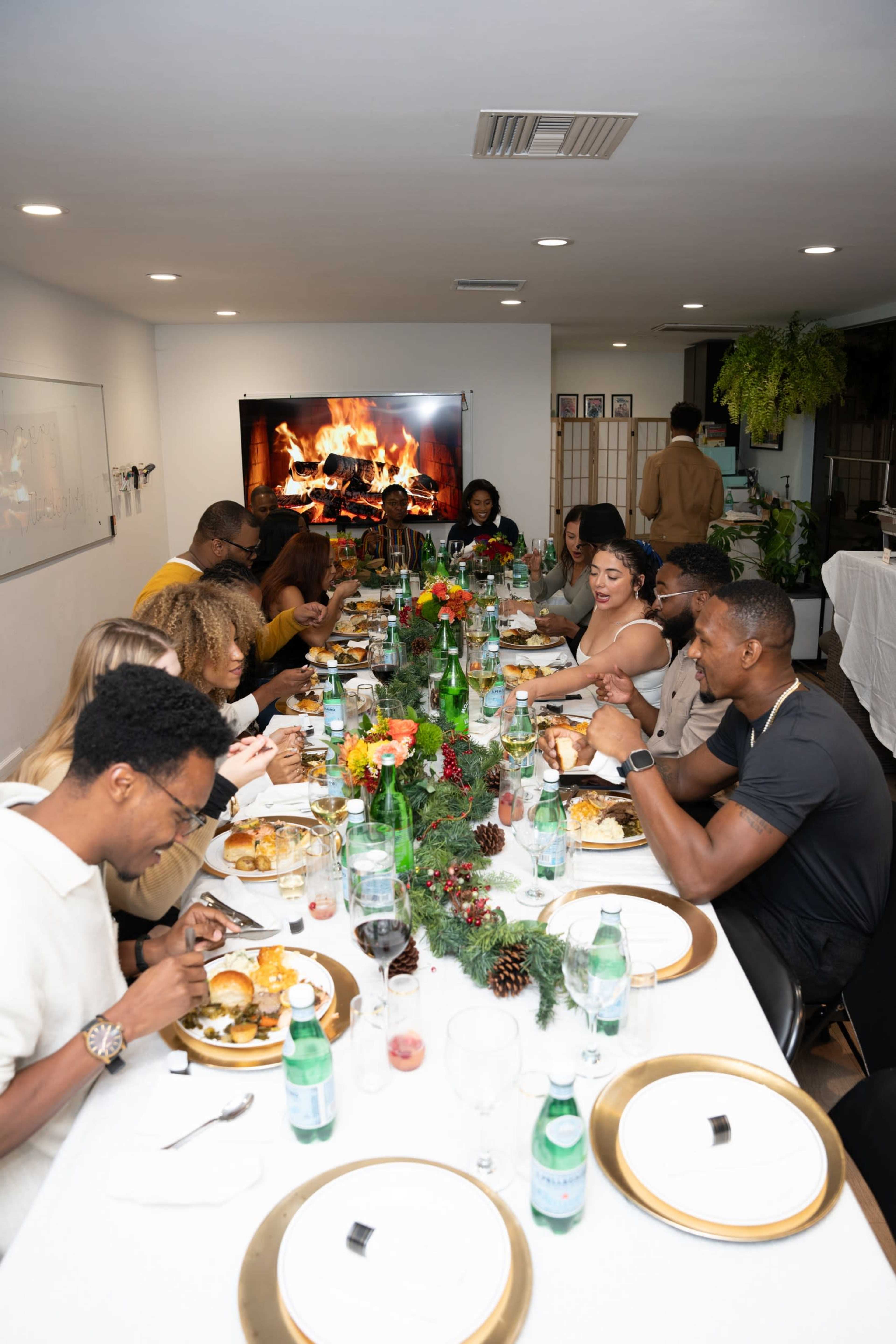 A large group of people is enjoying a festive dinner around a long table, with food and drinks in front of them and a fire visible on a screen in the background.