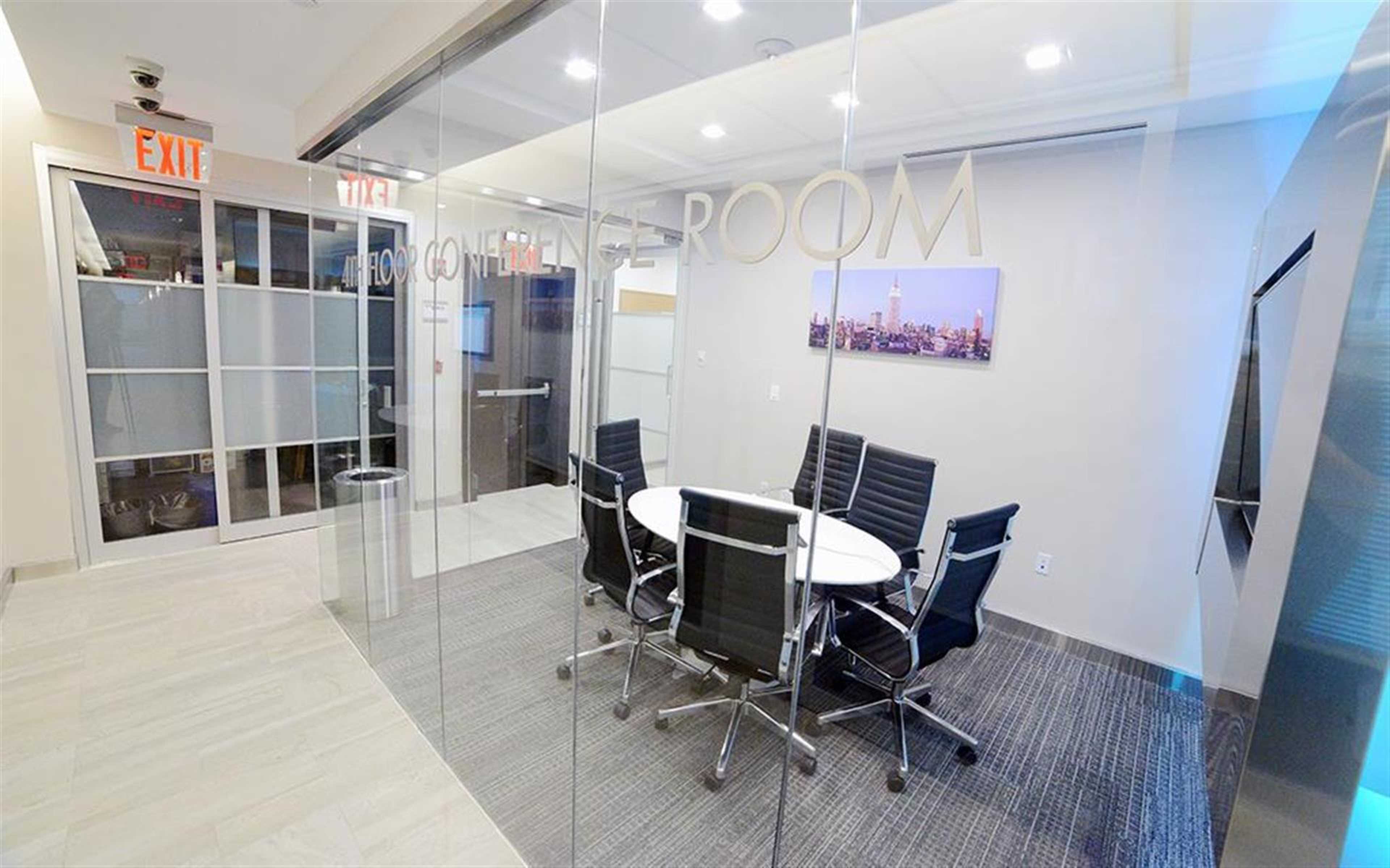 A modern conference room features a round table surrounded by black chairs, glass walls, and a large screen.