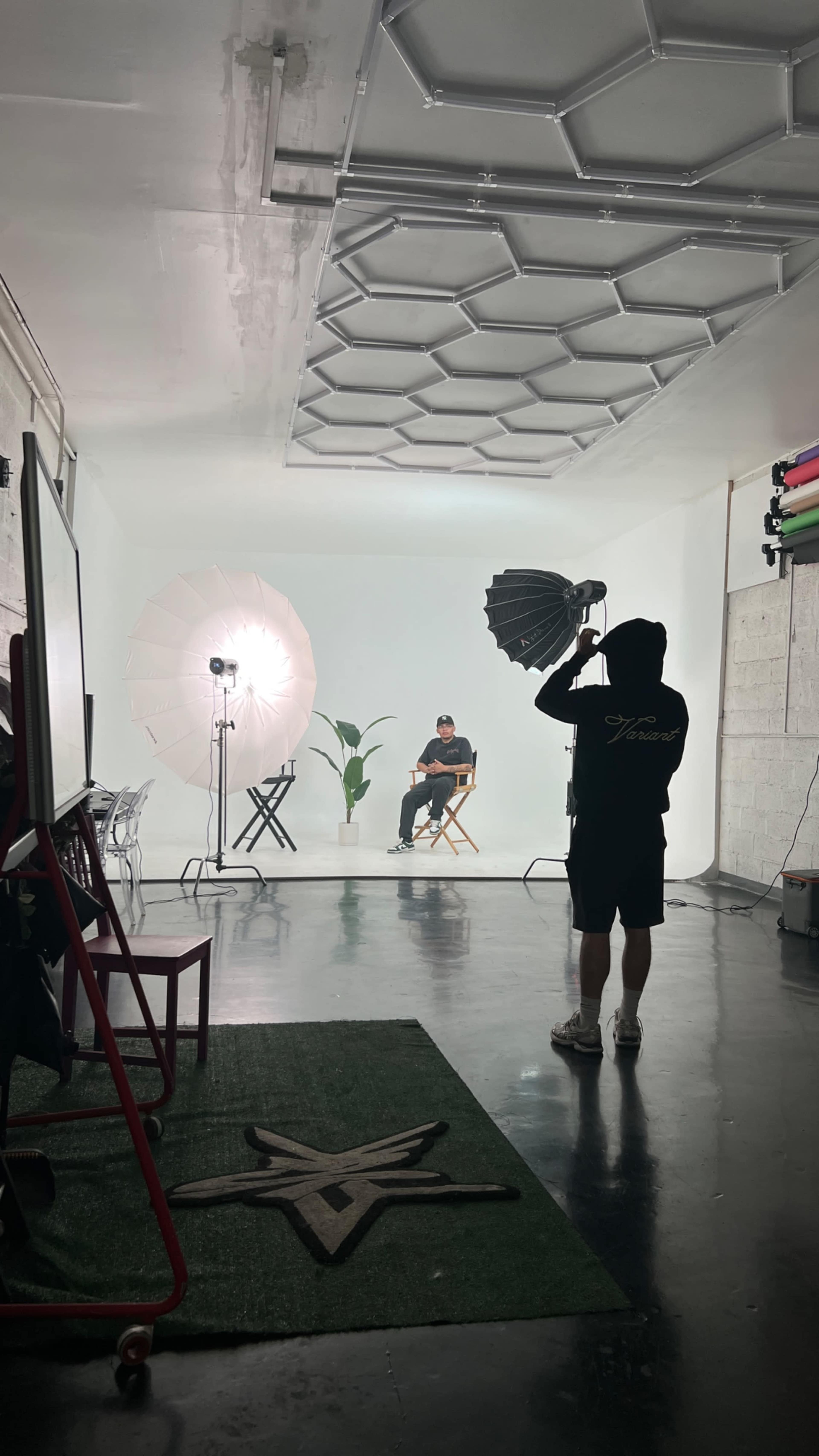 A photography studio features a seated subject under soft lighting, with various equipment set up including umbrellas, a chair, and a potted plant against a white background.