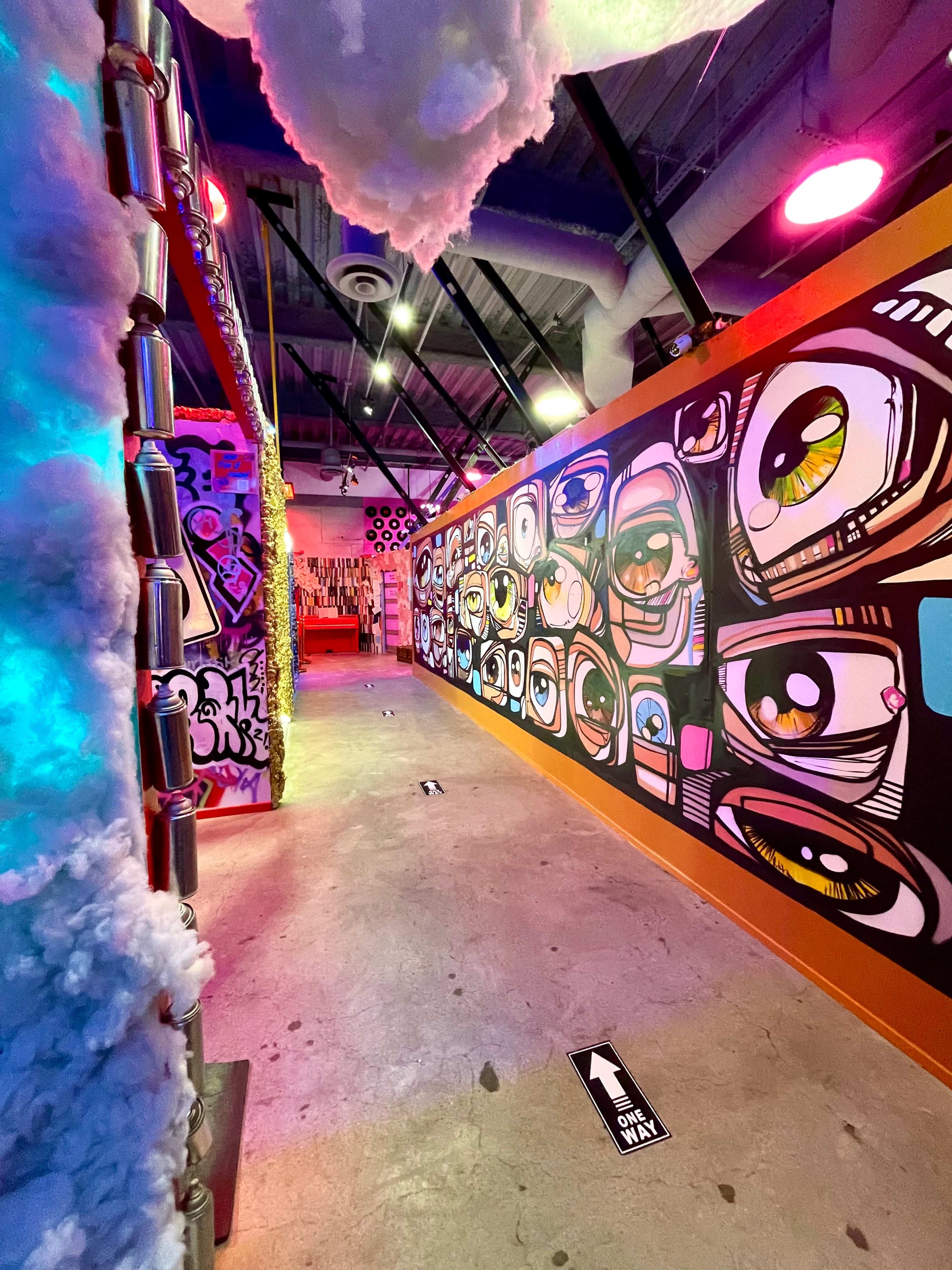 The scene features a brightly colored hallway adorned with large eye-themed murals and creative wall art, illuminated by vibrant lighting.