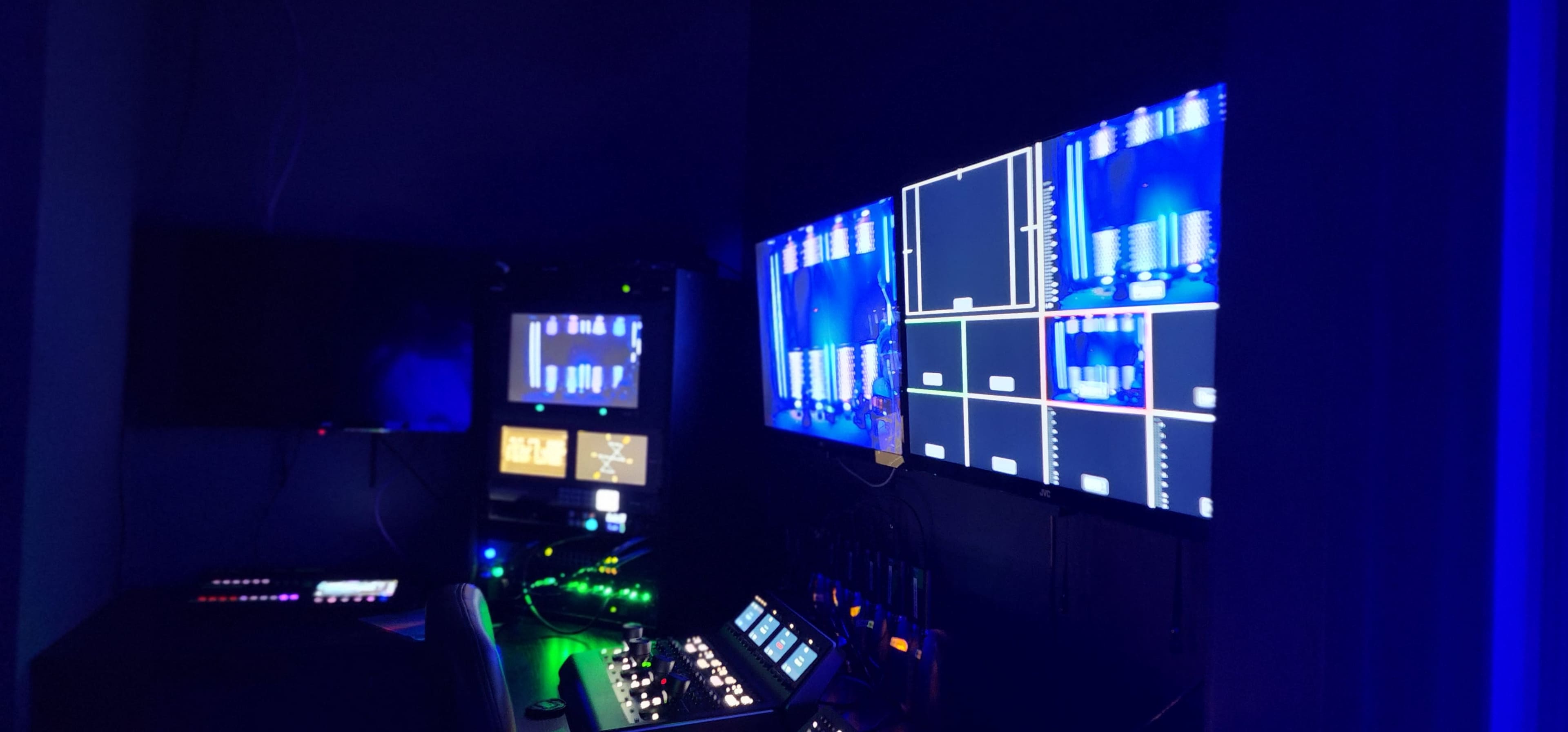 The image shows a dimly lit control room with multiple screens displaying various graphical outputs and digital controls.