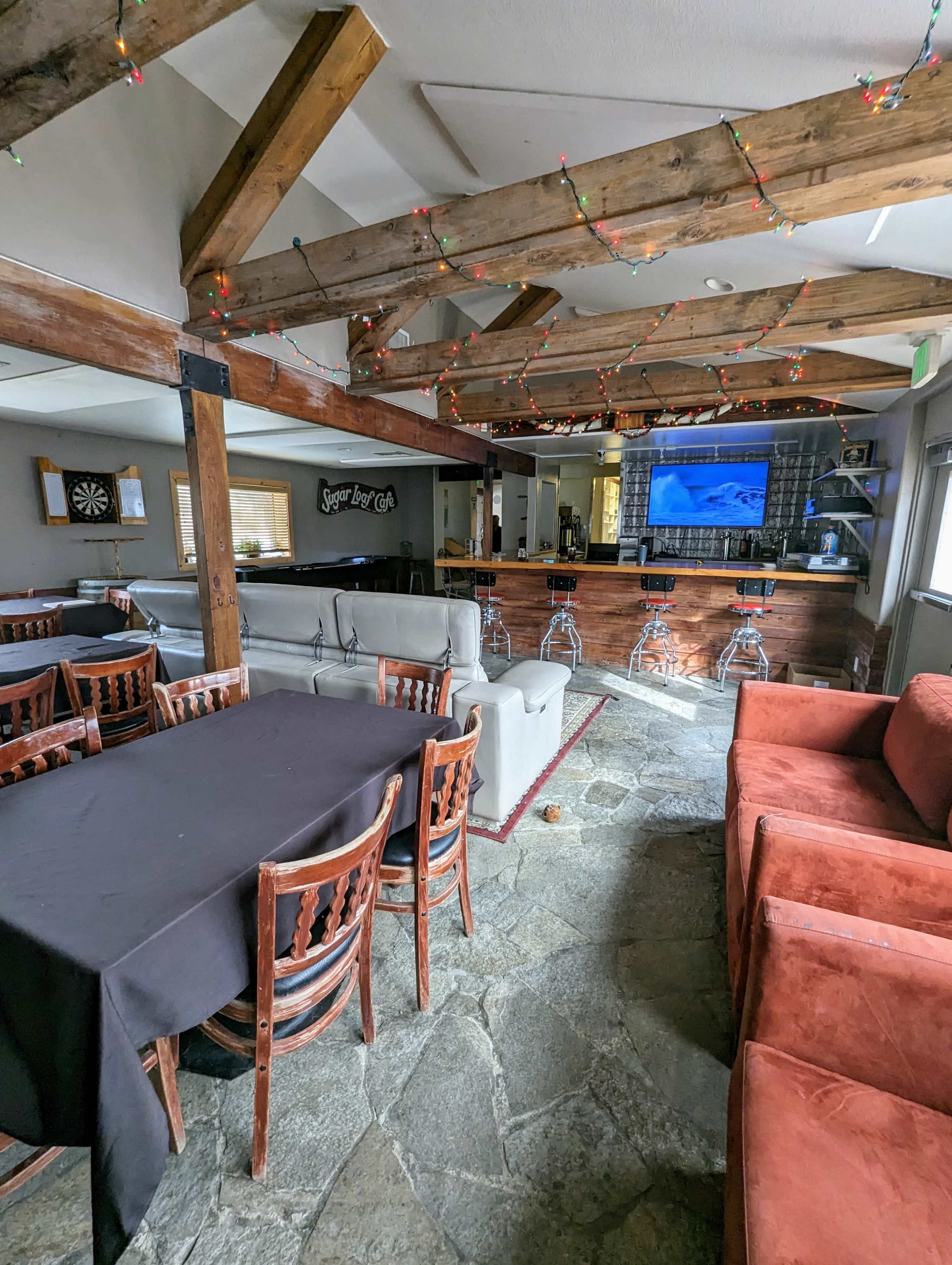 A spacious communal area with wooden beams, stone flooring, a bar with seating, and multiple tables arranged around a large screen.