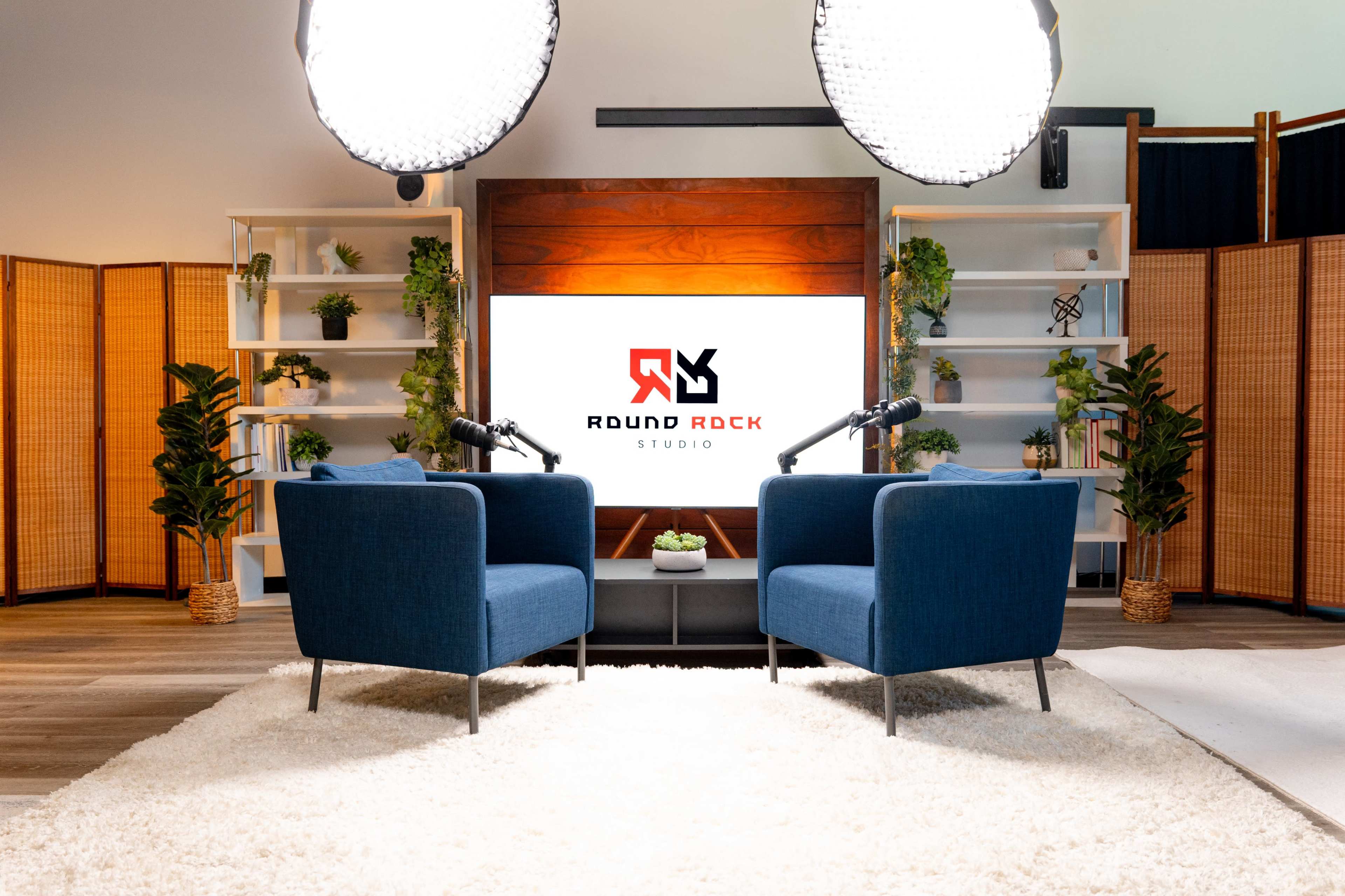 The image shows two blue armchairs facing a large screen with the "ROUND ROCK STUDIO" logo displayed, surrounded by plants and positioned under bright lights.
