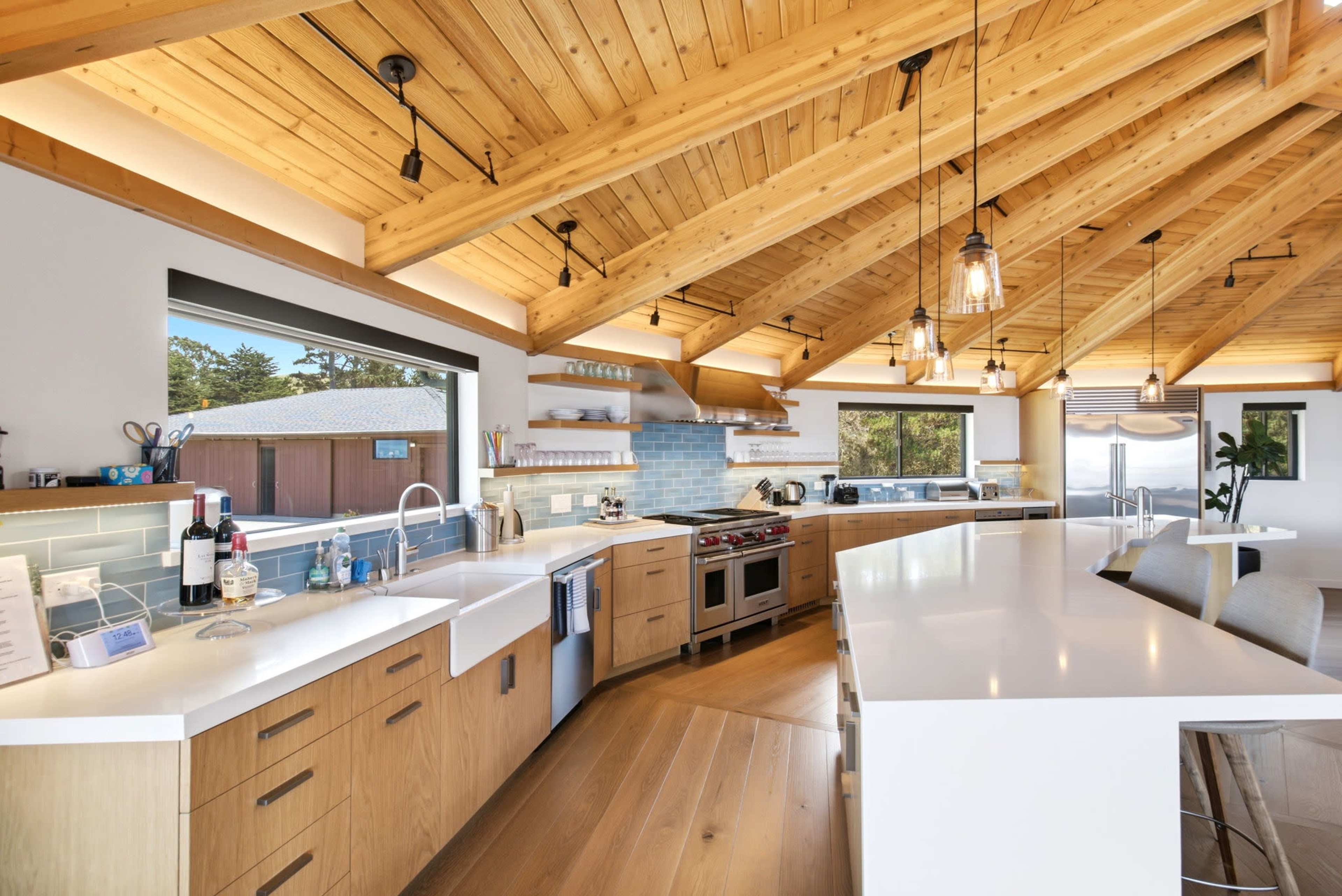 A modern kitchen features wooden beams, a large island, stainless steel appliances, and ample natural light from windows.