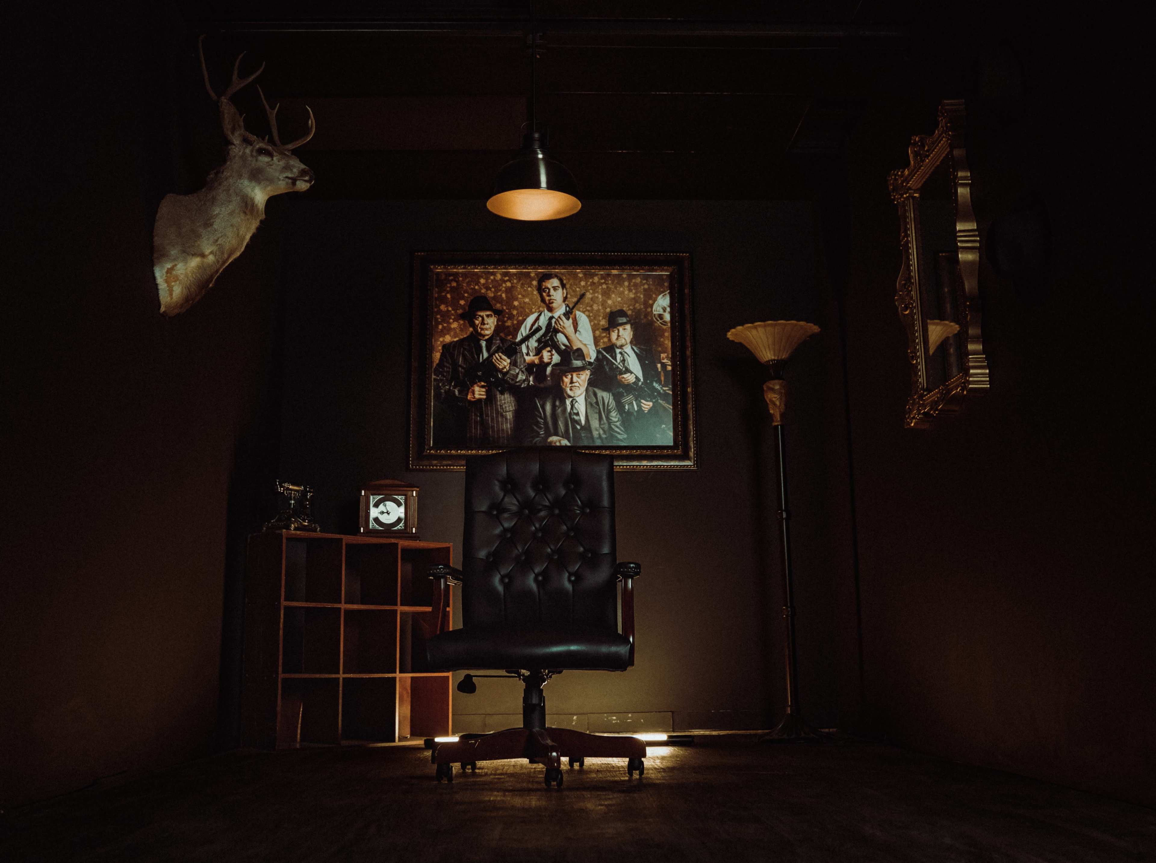 A dimly lit room with a black leather chair, a vintage painting of three men on the wall, a mounted deer head, and a lamp beside a wooden shelf.
