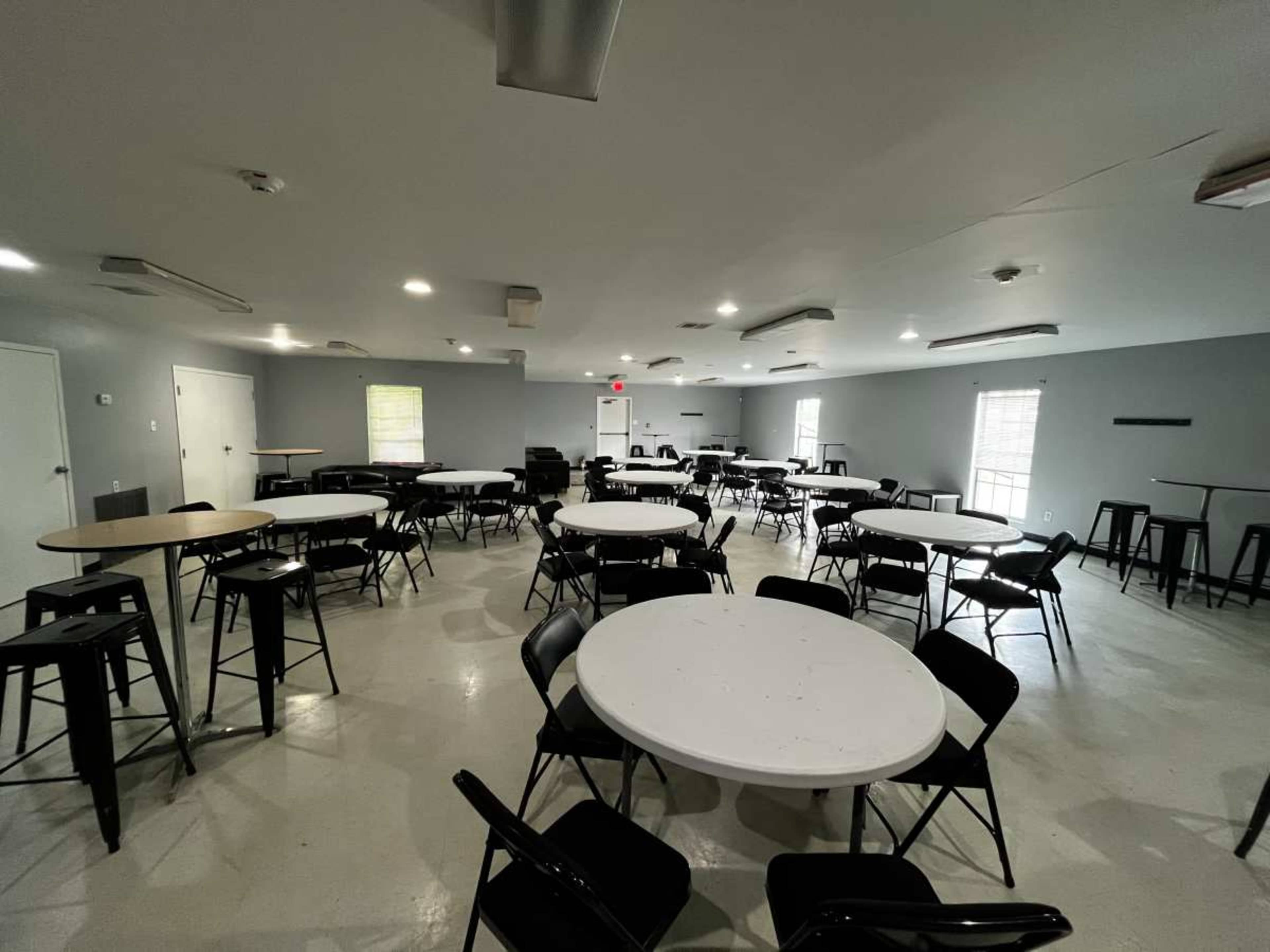 The image shows a spacious room containing several round and rectangular tables surrounded by chairs, with gray walls and bright overhead lighting.