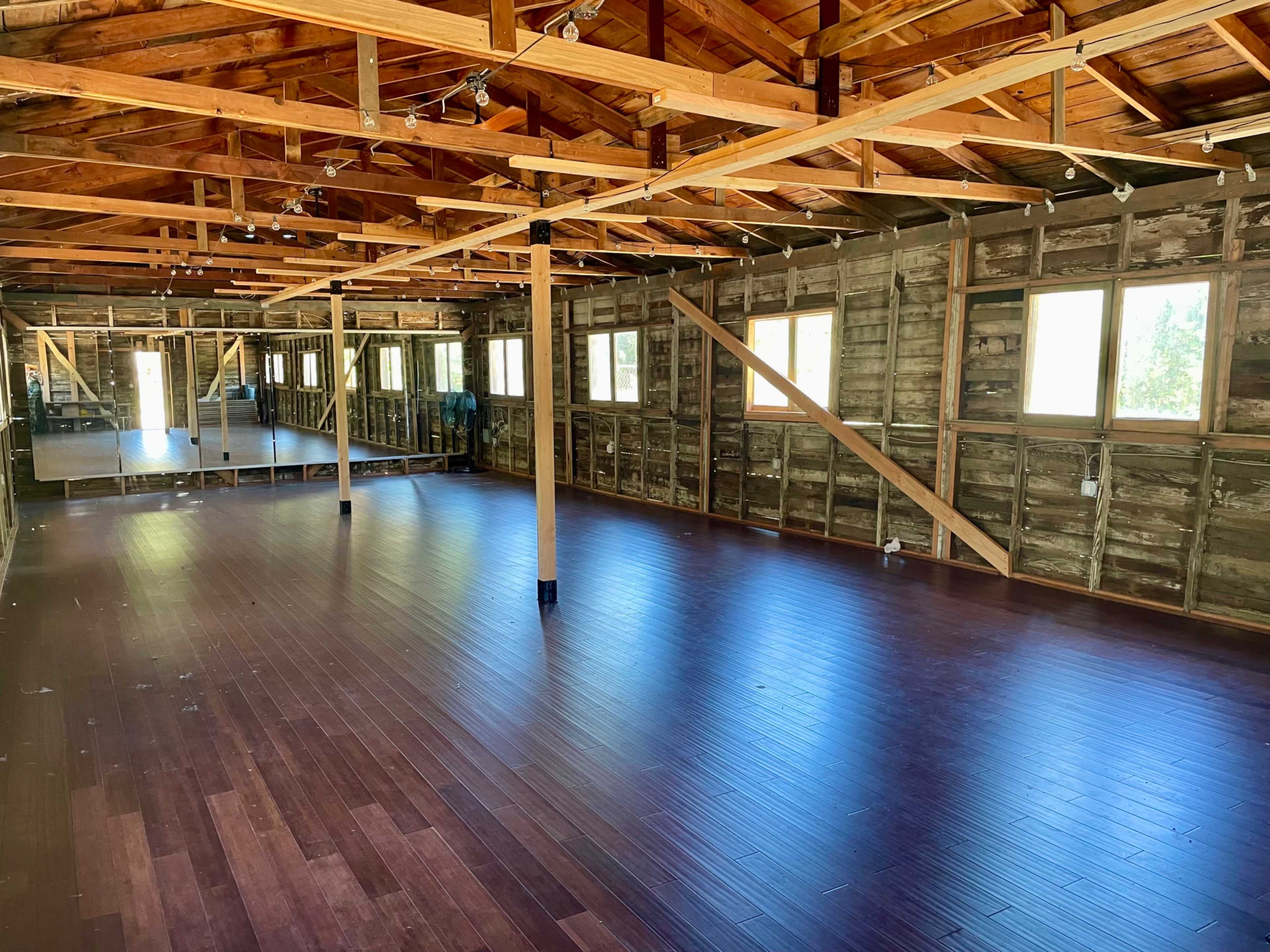 The image shows an empty room with wooden floors and exposed beams, featuring large windows and a mirrored wall on one side.