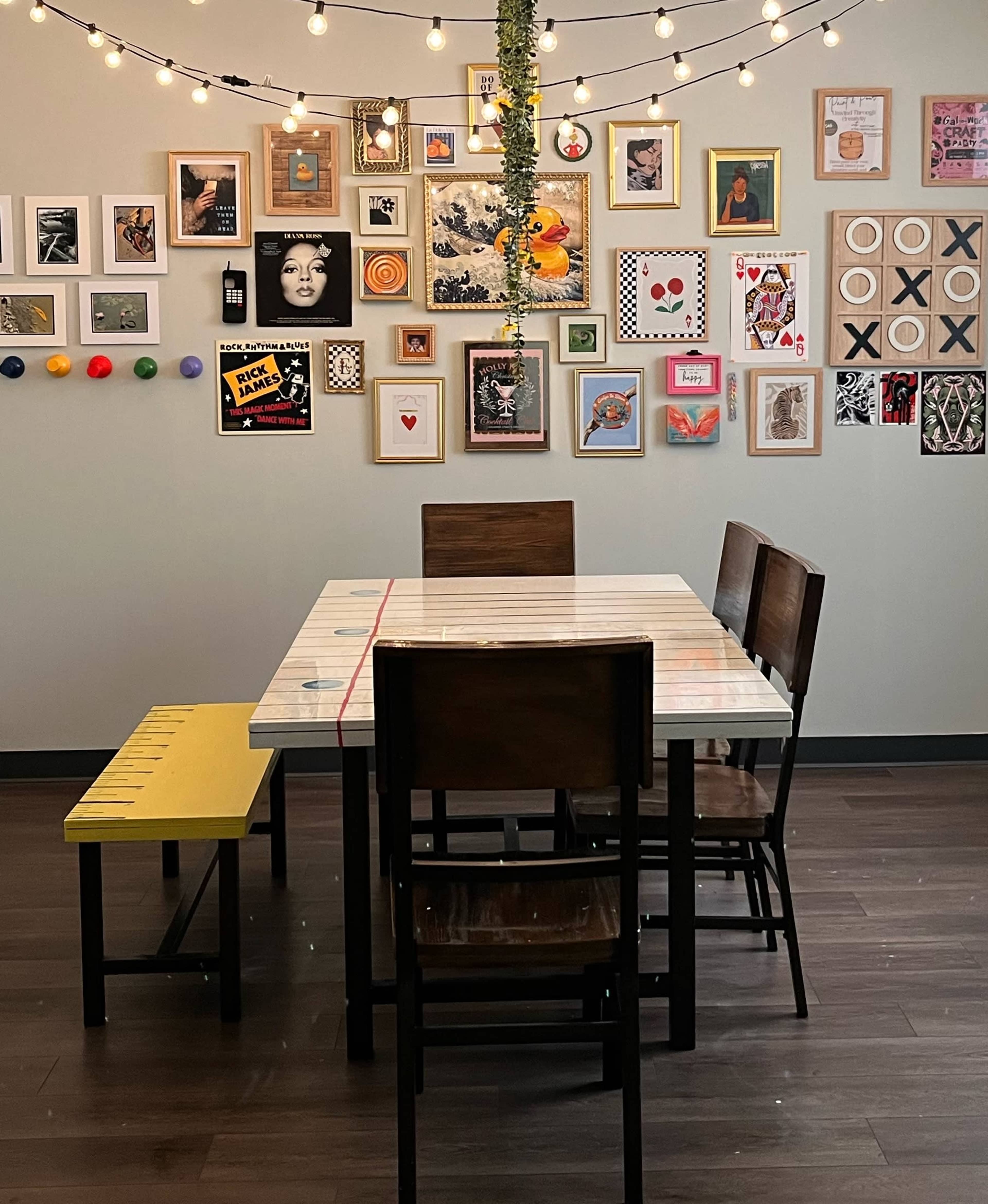 The image shows a dining area with a white table surrounded by four wooden chairs and a yellow bench, set against a wall decorated with various framed artworks and string lights.