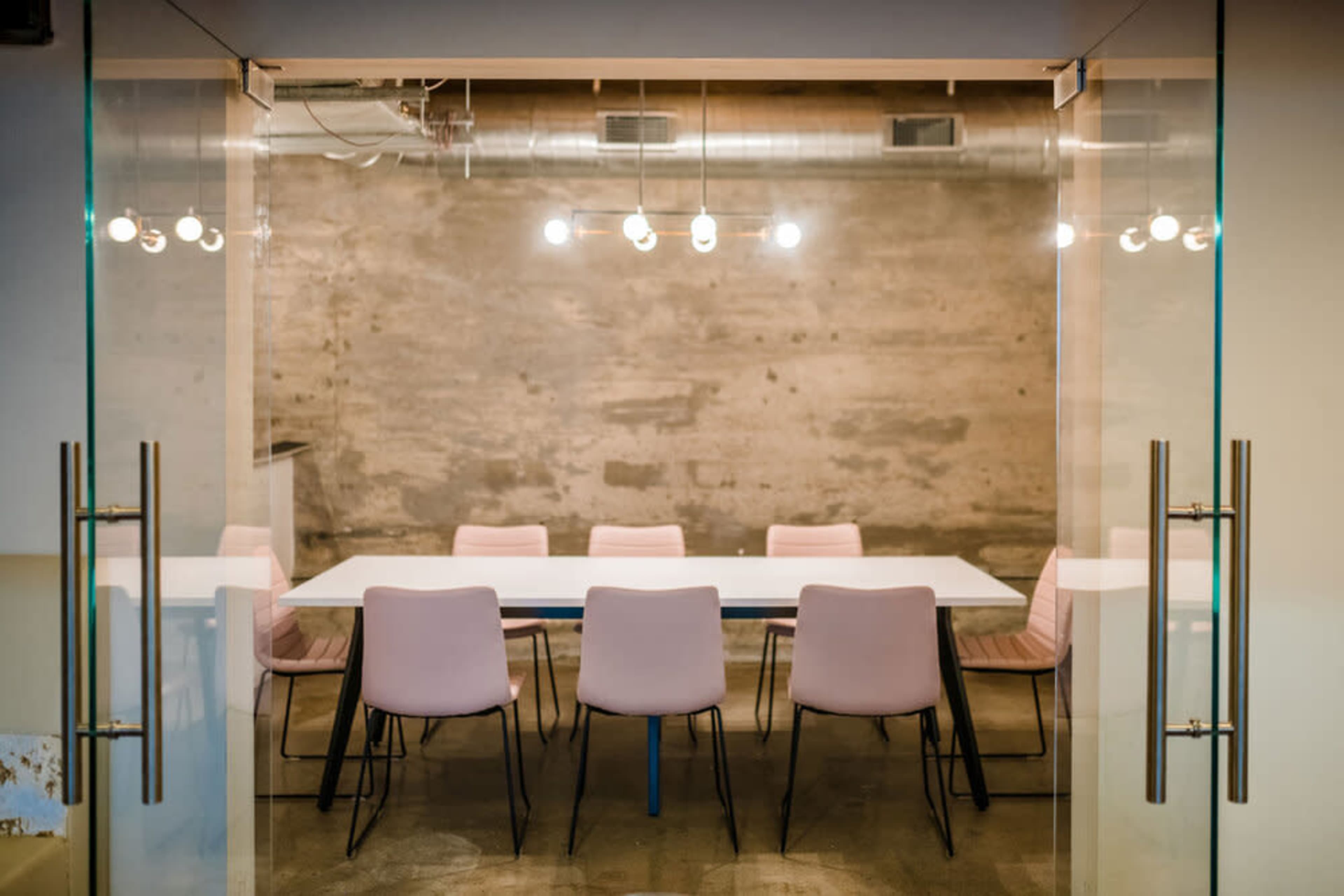 A minimalist conference room features a long white table surrounded by pink chairs under industrial-style lights.