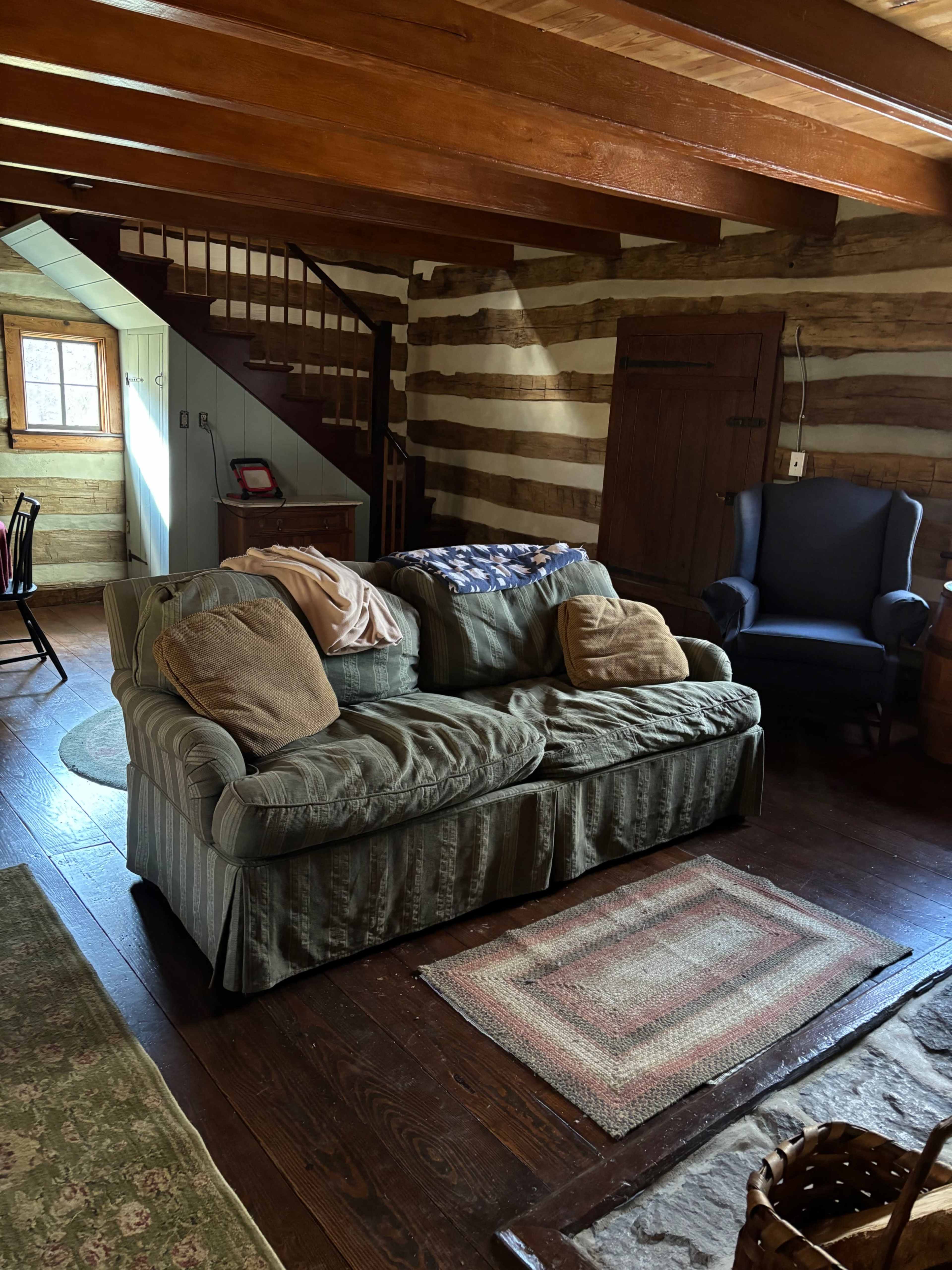 The image shows a cozy, rustic living room with a couch and chairs surrounded by wooden walls and a staircase in the background.