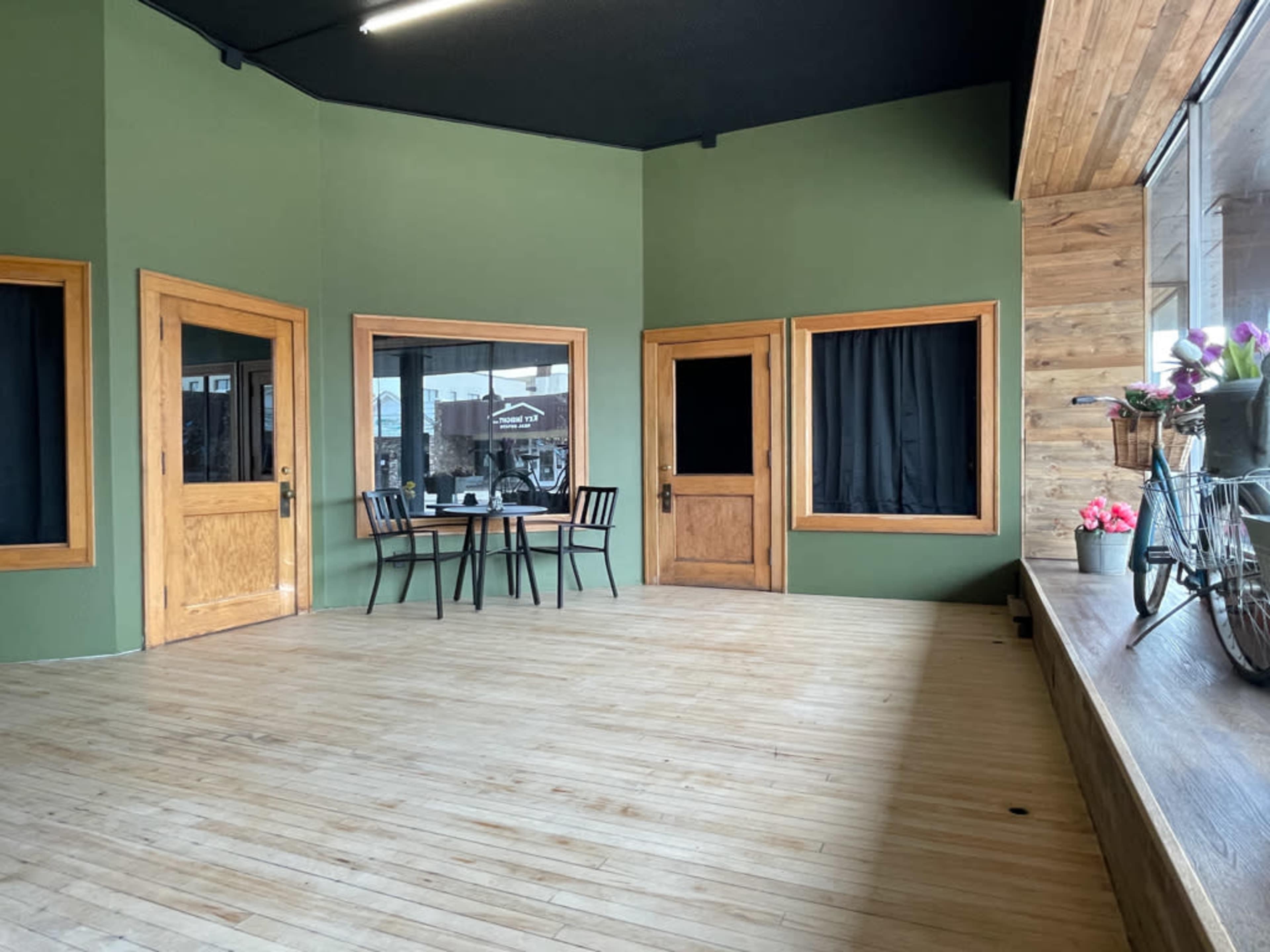 A spacious room with green walls, hardwood flooring, two wooden doors, a small table with two chairs, and large windows displaying a view of the outside.