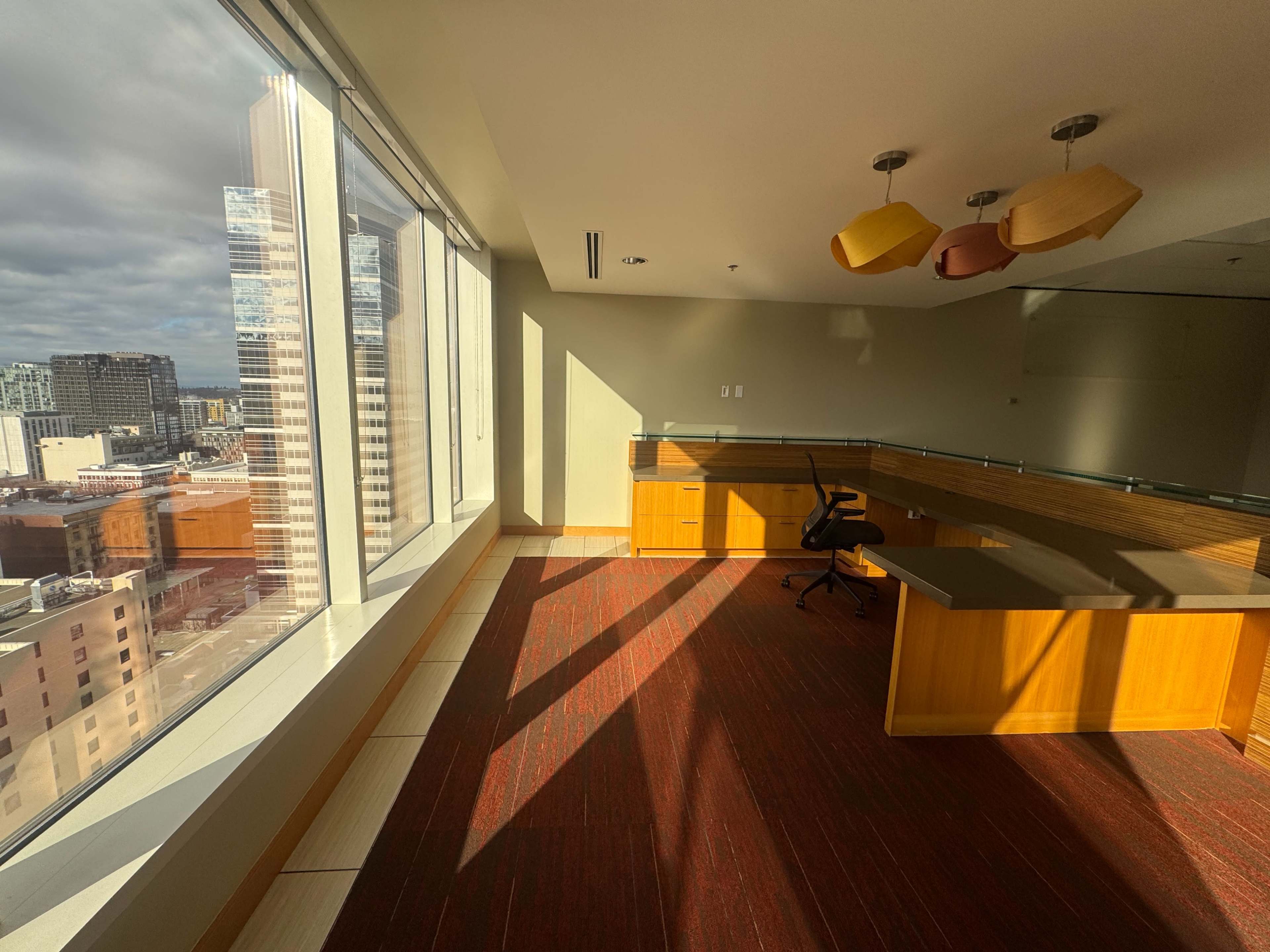 An office space with large windows showcasing a city skyline, featuring wooden furniture and a black office chair.