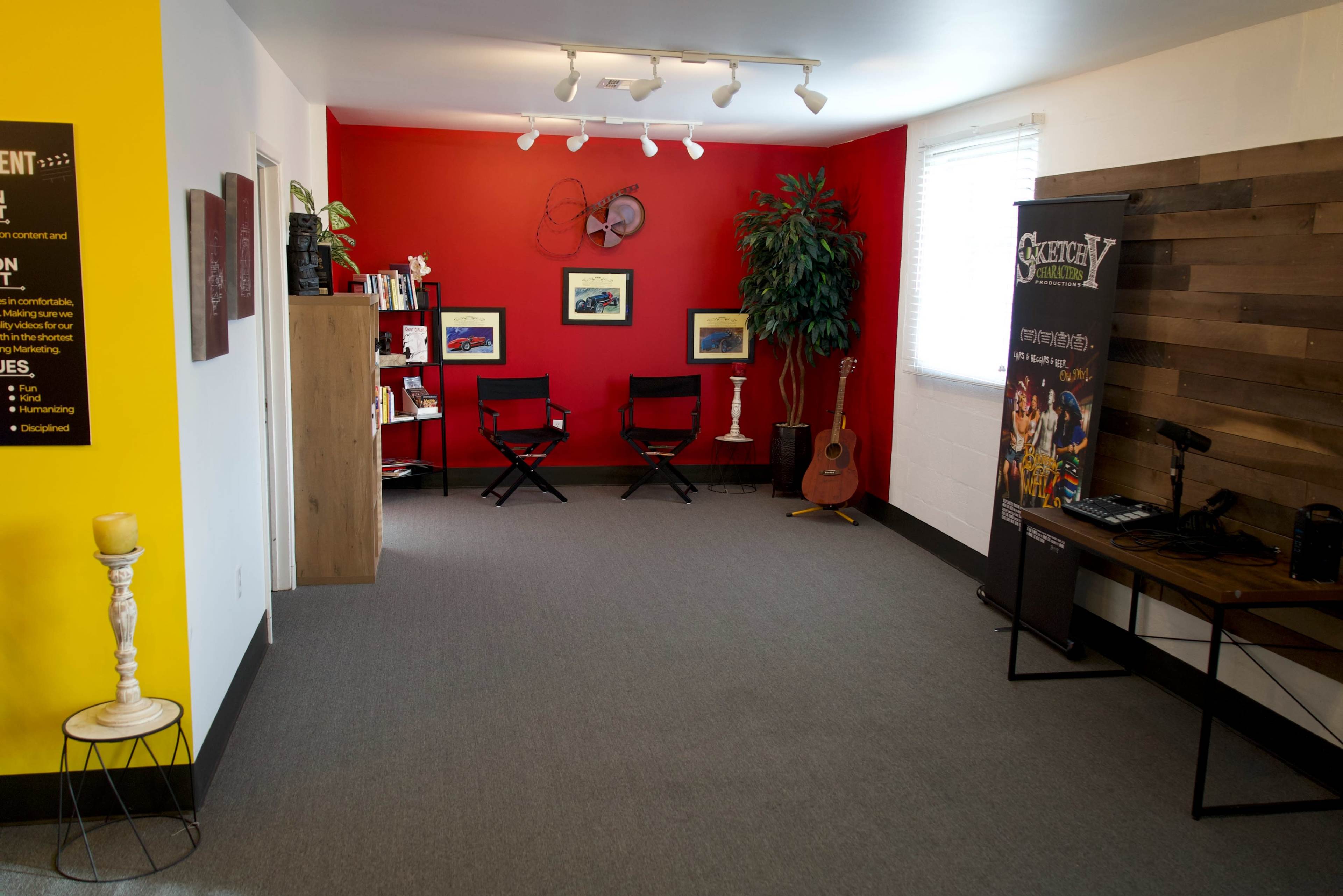A well-lit room with a red accent wall, featuring two black director's chairs, a wooden shelf with books, a guitar, and a promotional banner.