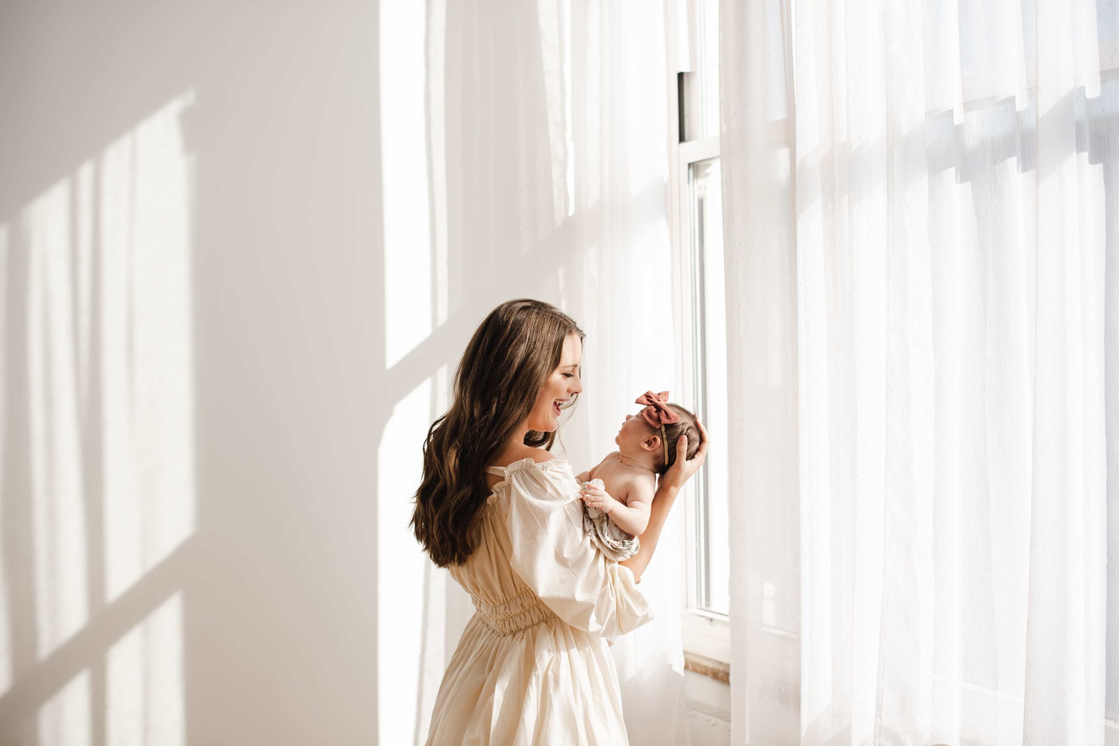 A woman holds an infant while standing near a window with sheer white curtains.