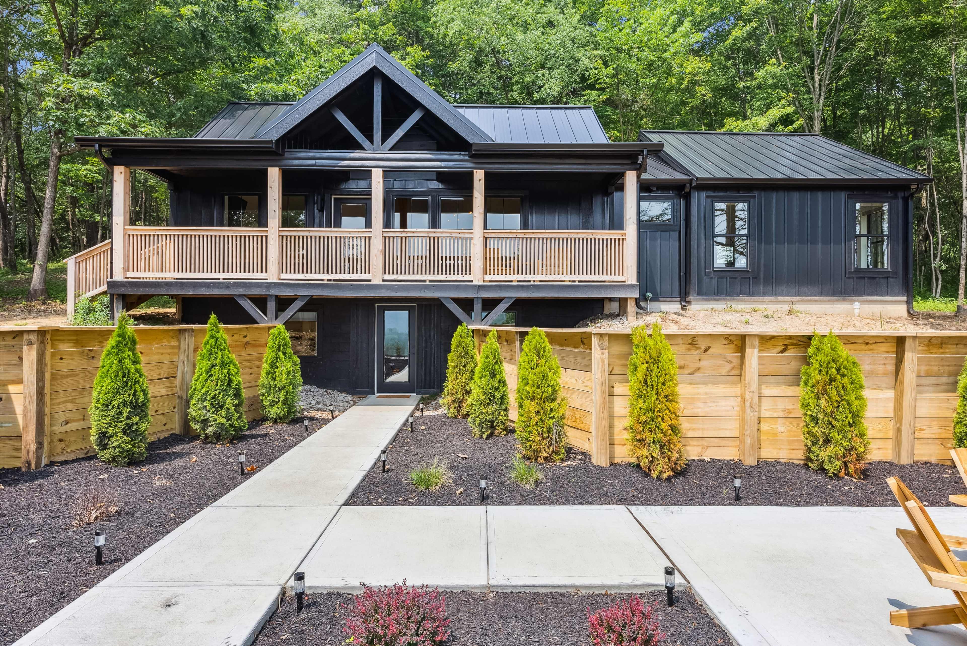 A modern black house with a balcony, surrounded by neatly arranged shrubs and a concrete pathway leading to the entrance.