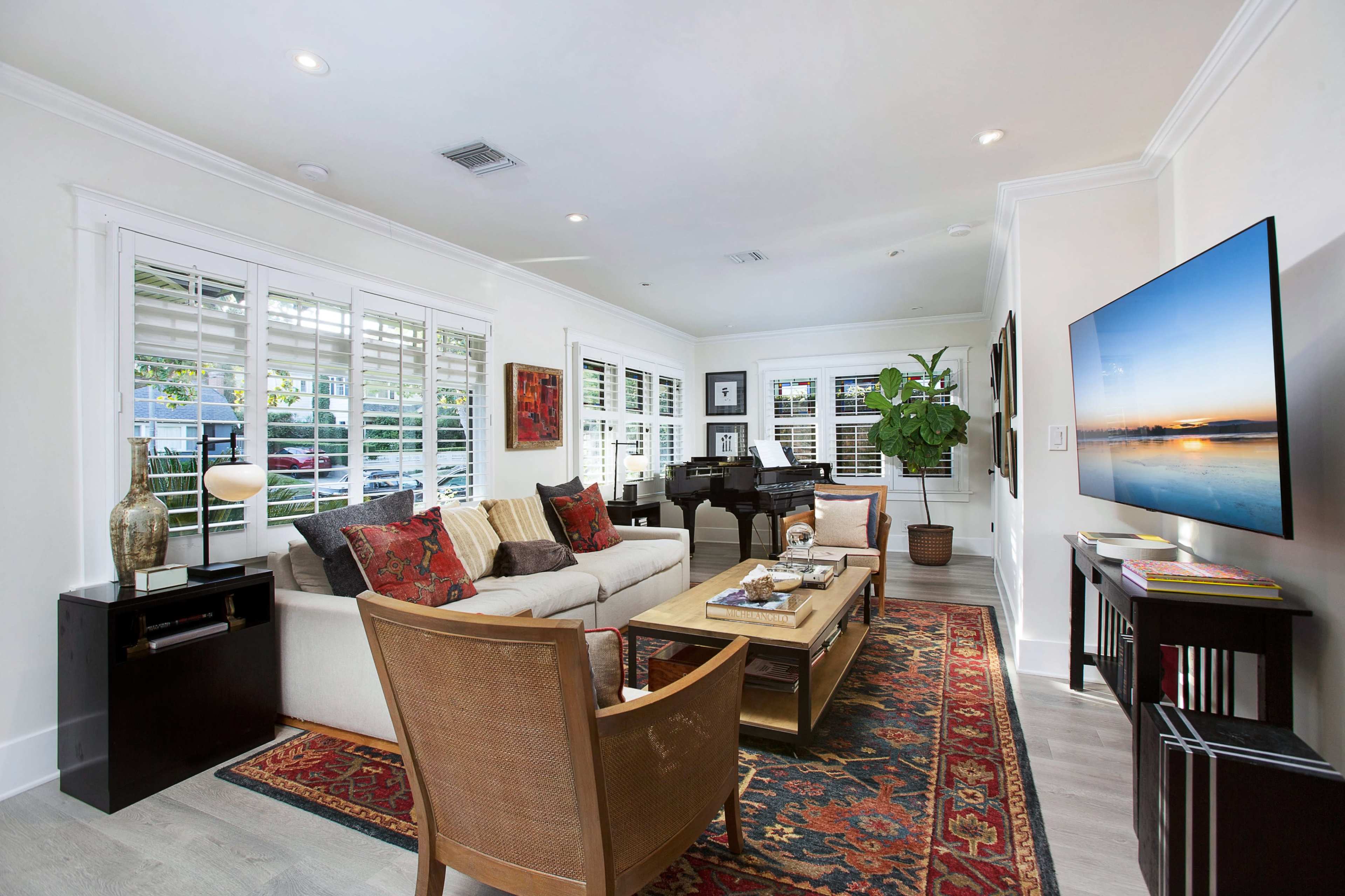 The living room features a sofa, armchairs, a coffee table, and a television, with natural light filtering through window shutters.