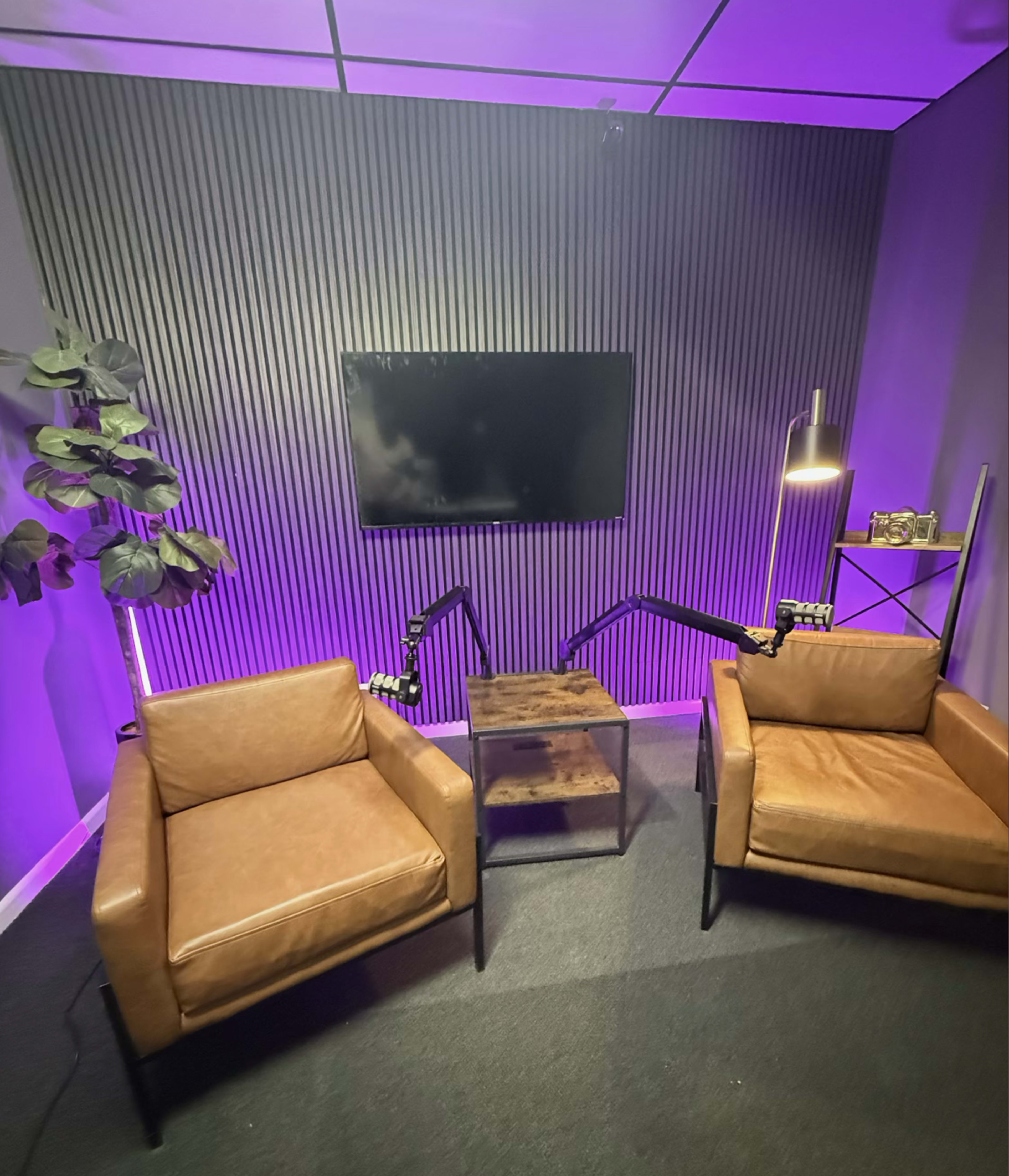 The image shows a modern conversation space featuring two brown leather chairs, a small table, a wall-mounted TV, and accent lighting in a purple hue.
