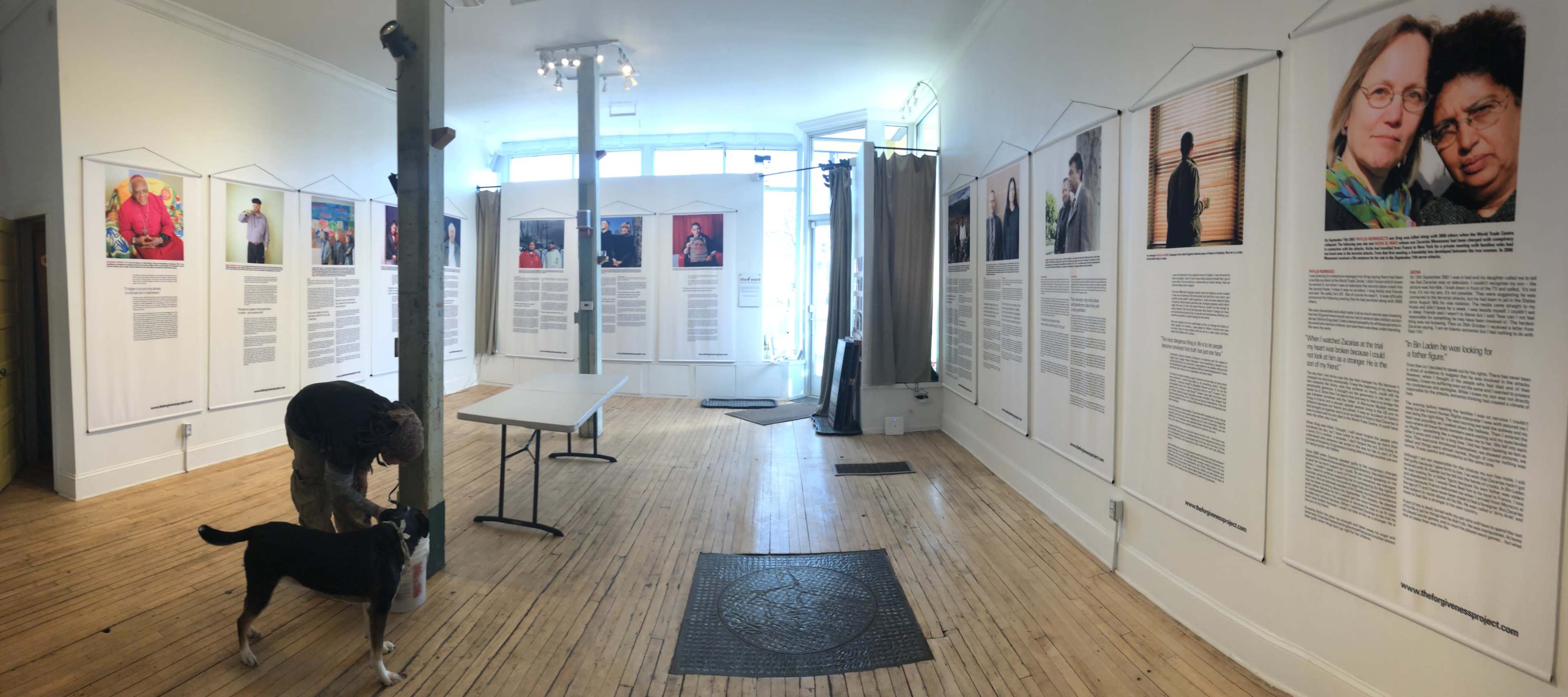 An indoor gallery space with several large framed photographs and text panels displayed on the walls, featuring a person with a dog near a table in the center.