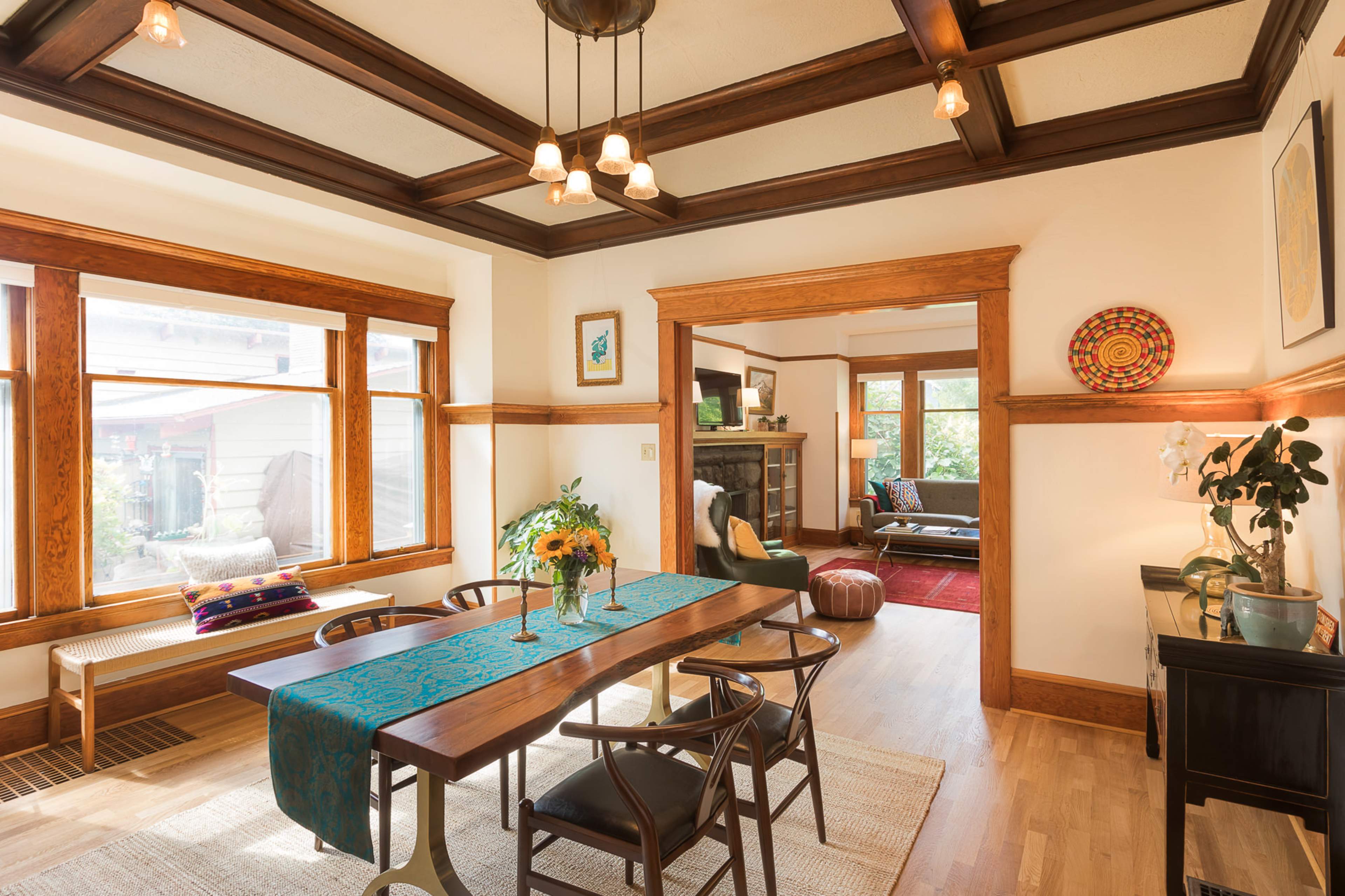 A dining area with a wooden table set for a meal, surrounded by chairs, featuring large windows that let in natural light and an open doorway leading to a living room.