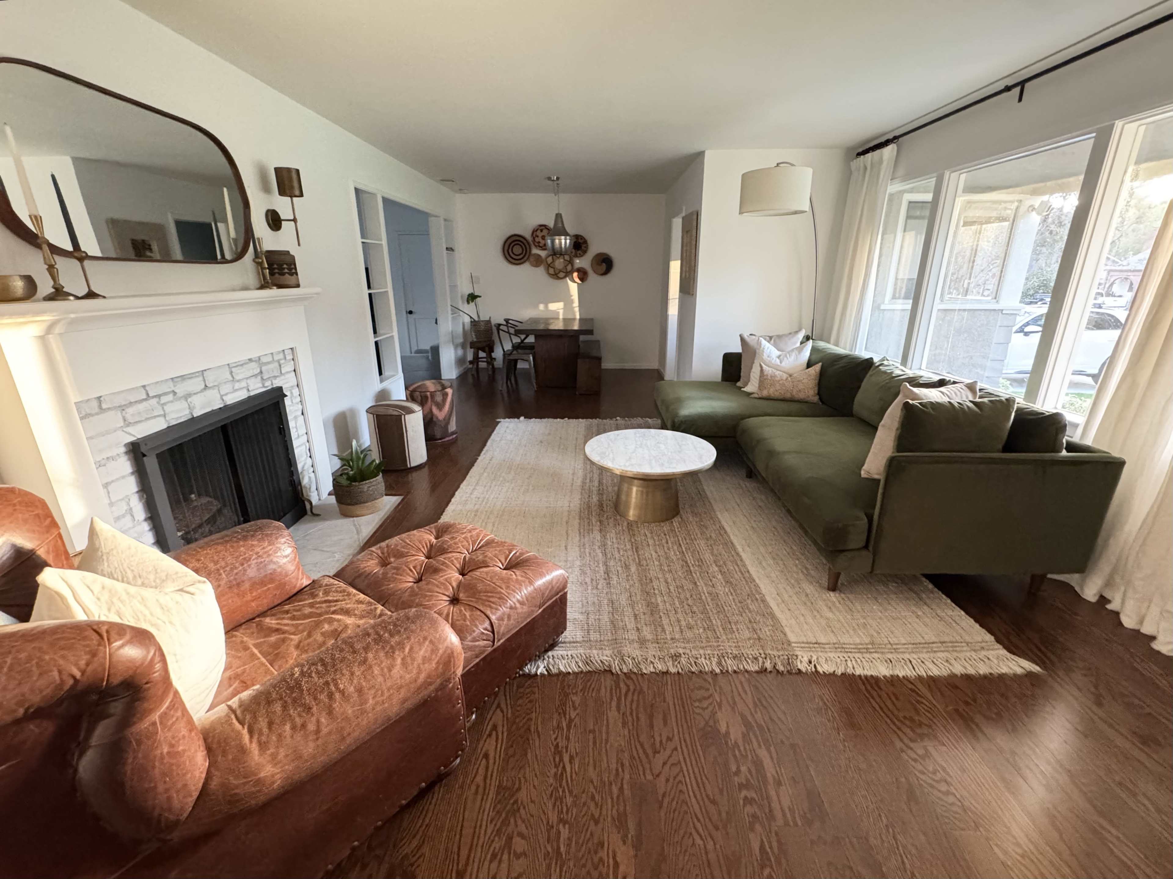 The living room features a green sectional sofa, a brown leather chair, a round coffee table, and a fireplace, with natural light coming through large windows.