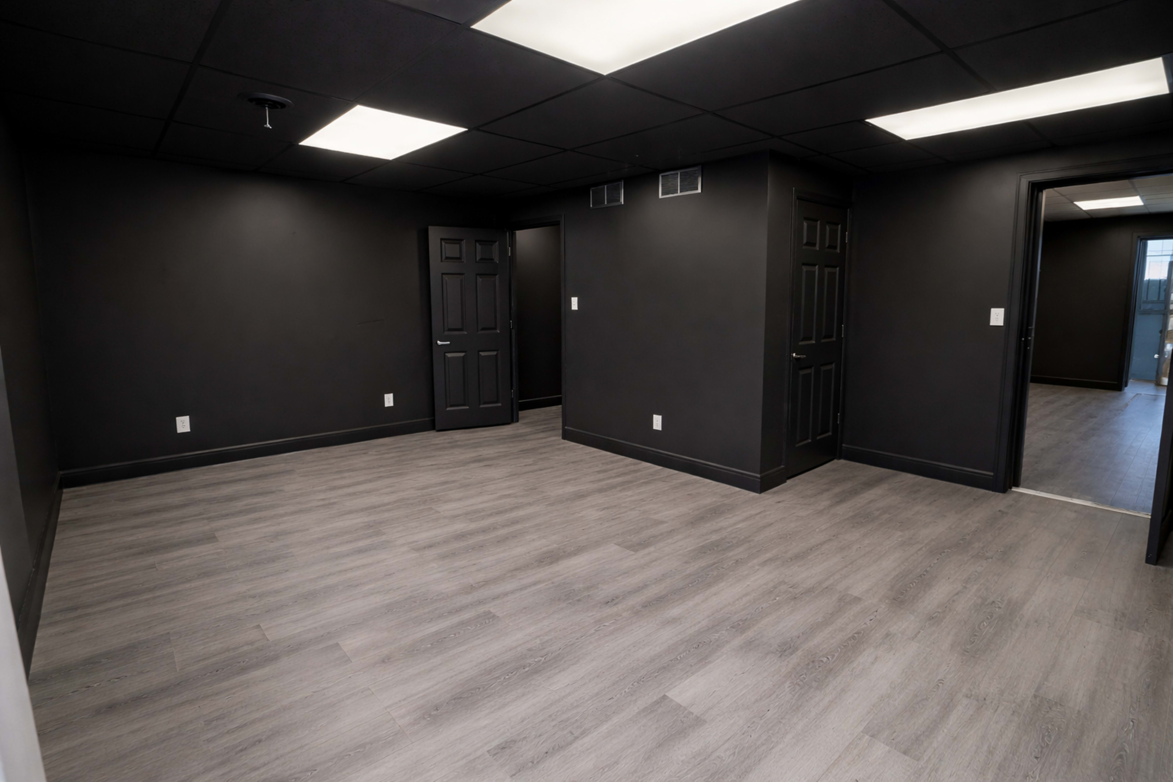 The image shows an empty room with black walls, a gray laminate floor, and multiple doors leading to adjacent areas.