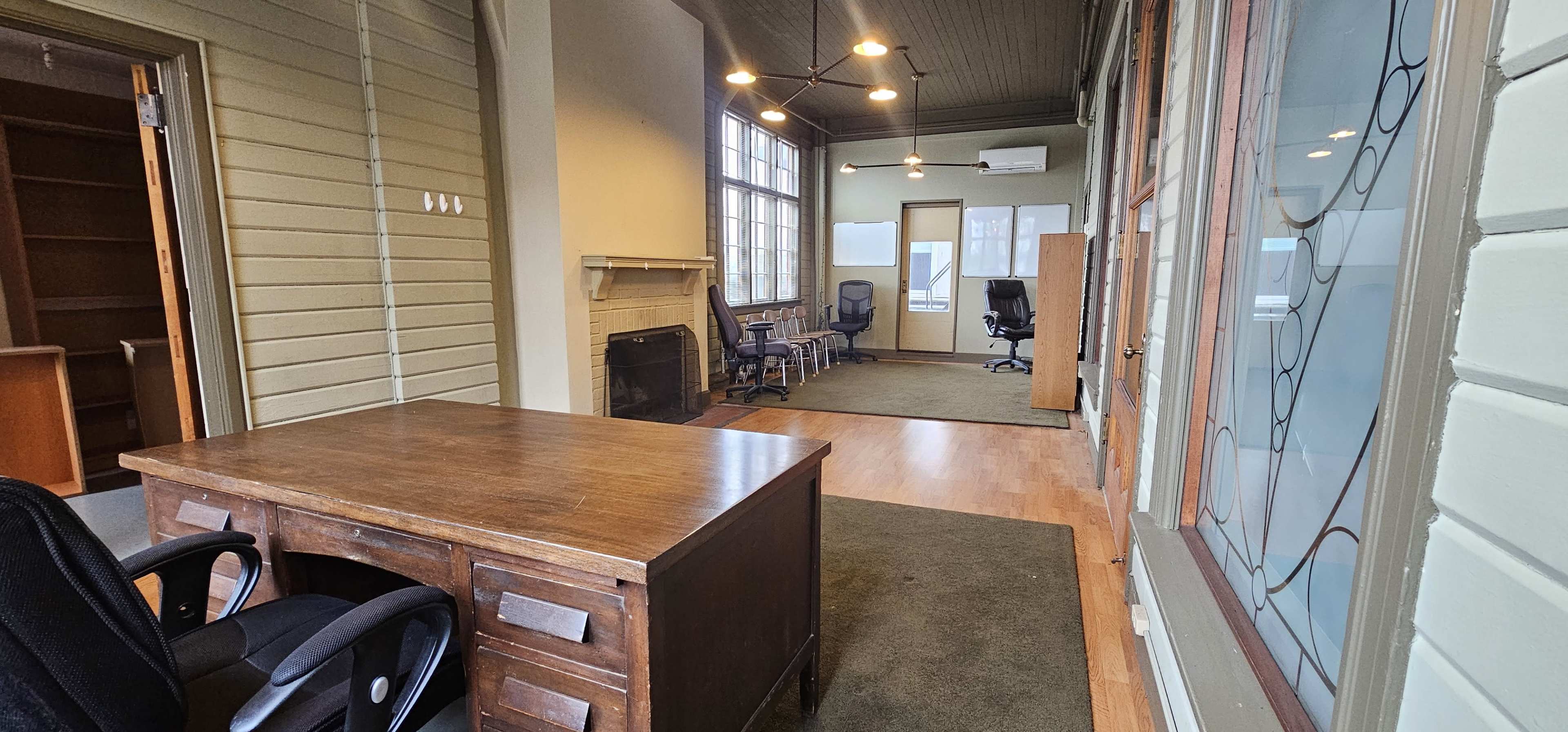 An office space with a wooden desk, several chairs, a fireplace, and large windows allowing natural light.