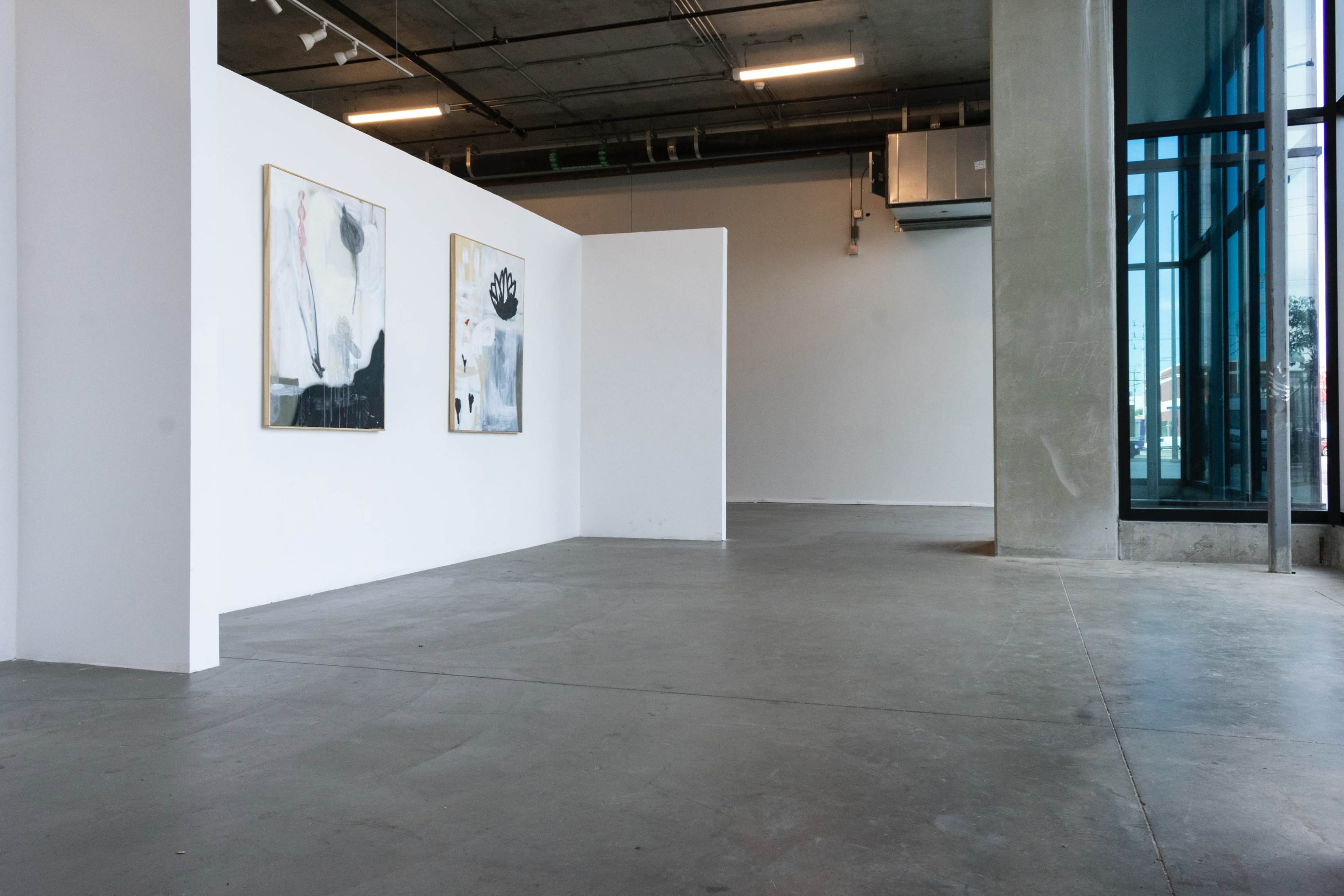 A spacious interior of an art gallery featuring two abstract paintings on a white wall and large windows allowing natural light to enter.